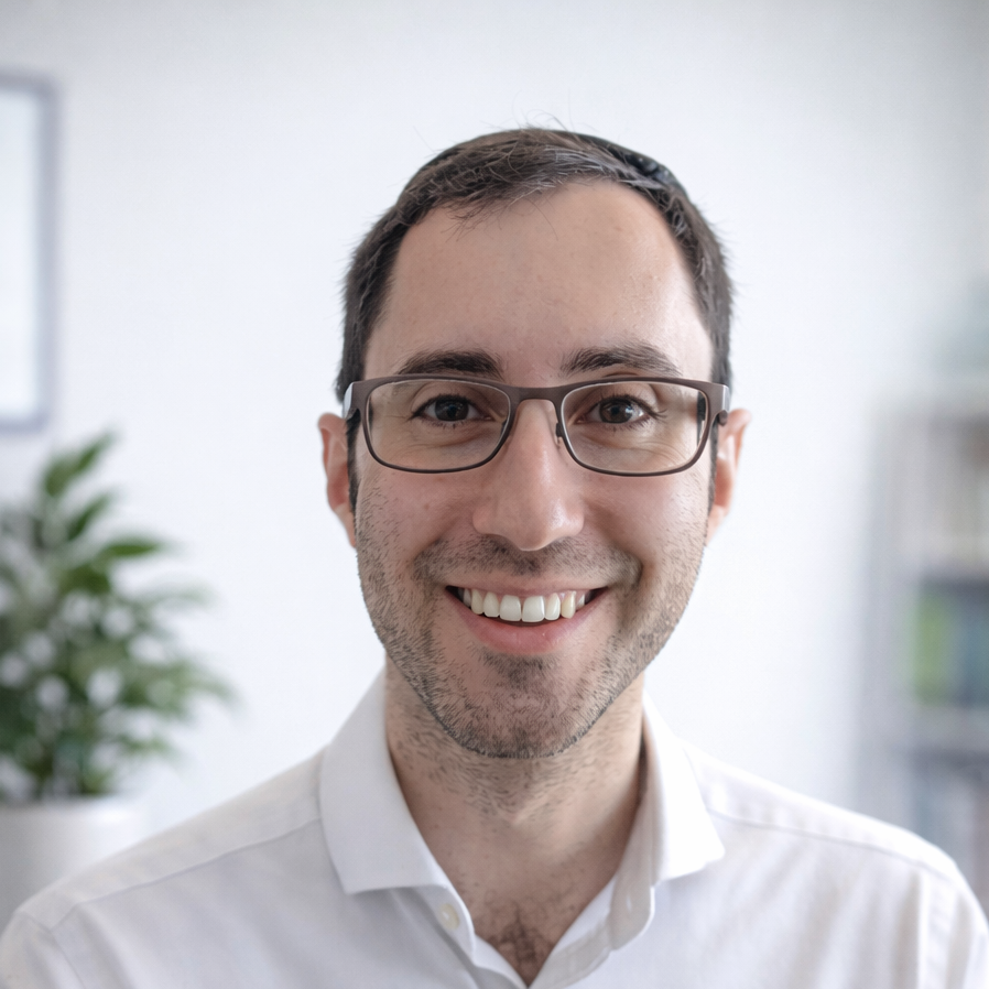 A smiling man with glasses, dark hair, and light skin, wearing a white shirt, indoors with a blurred plant and bookshelf in the background. the lead psychiatrist in our adhd, autism and general psychiatry team.