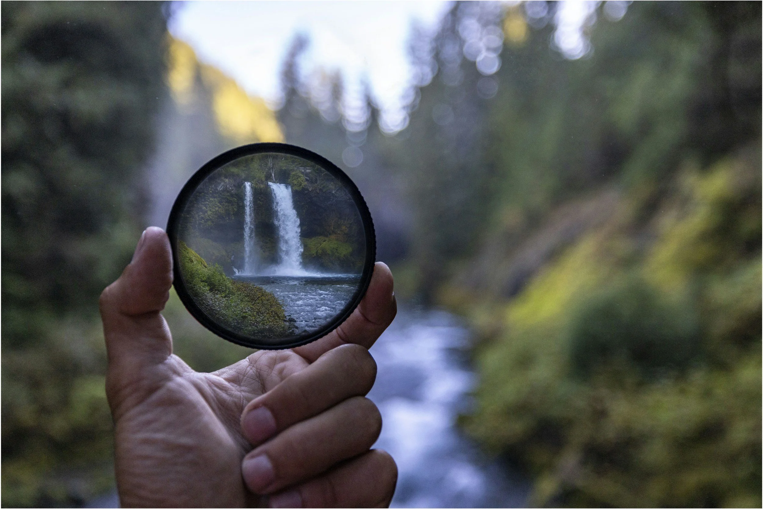 Person holding a circular mirror reflecting a waterfall in a forest representing transparent and affordable prices for ADHD and psychiatric treatment.
