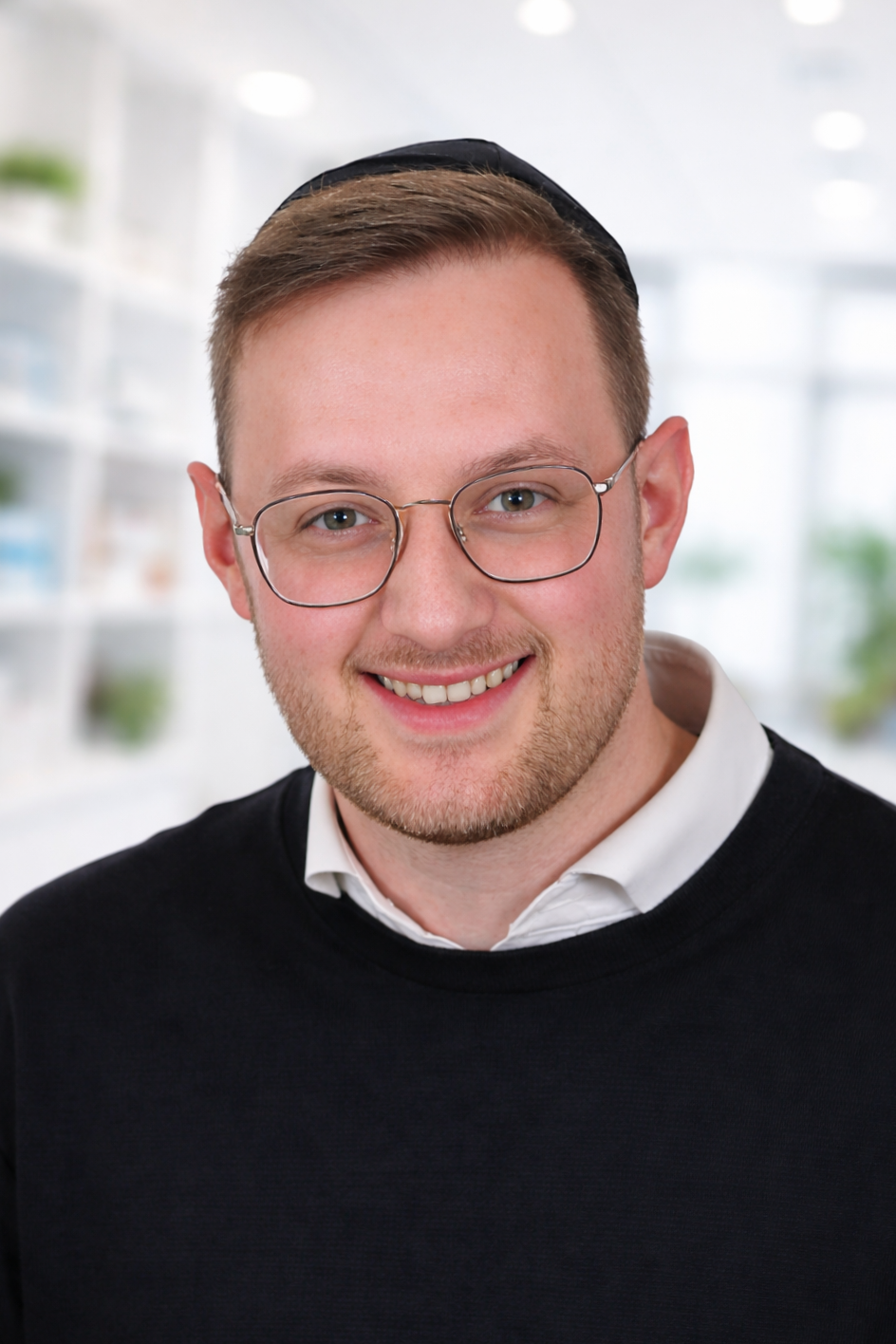 A young man wearing glasses, a white shirt, a black sweater, and a black kippah smiling at the camera, with a blurred bright indoor background. an assistant mental health practitioner in our adhd, autism and general psychiatry team.