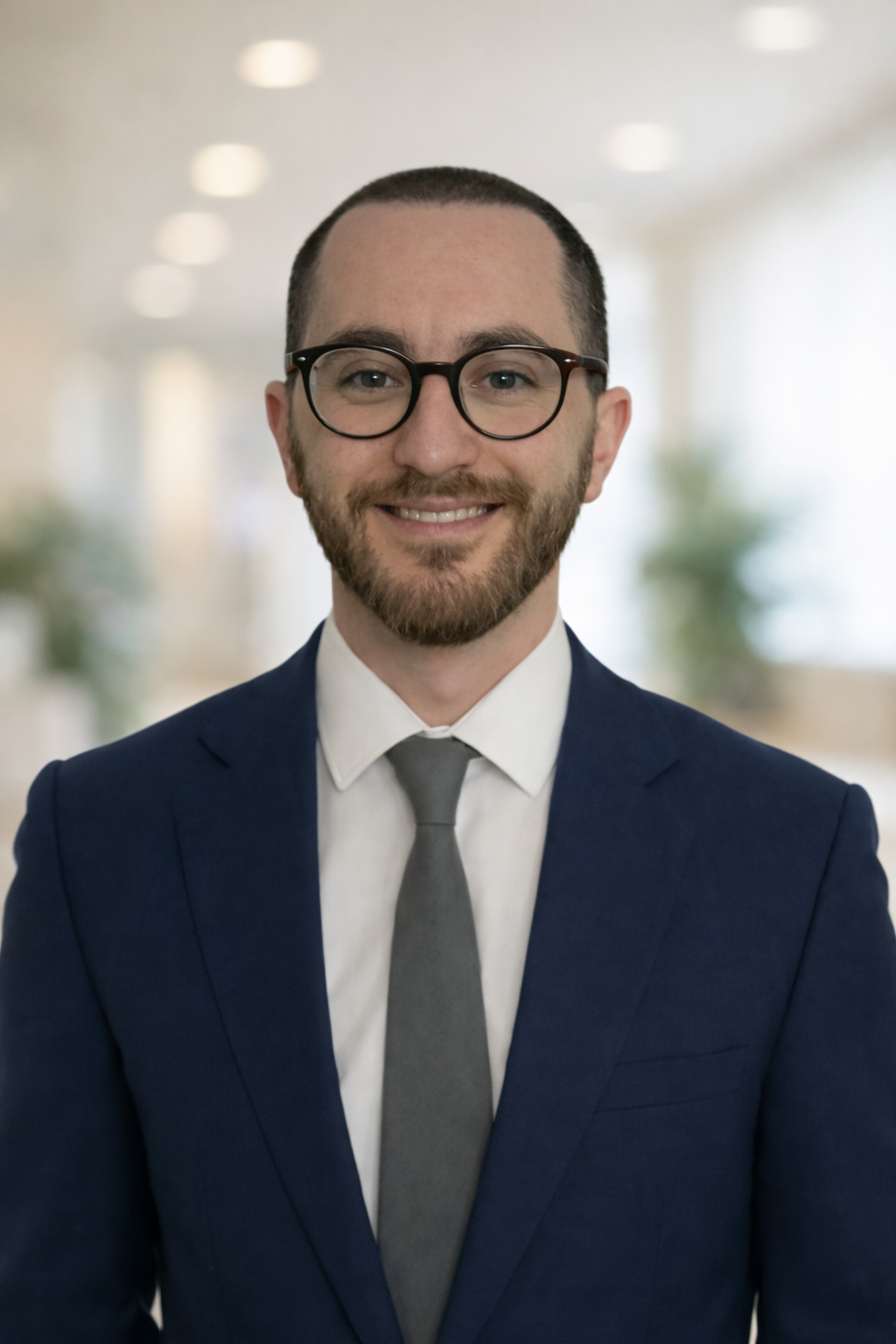 A man in a navy blue suit, white shirt, and gray tie, smiling with glasses and a beard, standing in a well-lit indoor space. a resident doctor in our adhd, autism and general psychiatry team.