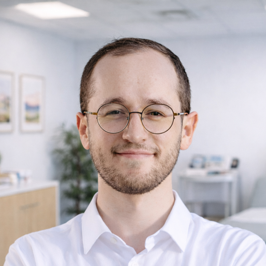 Portrait of a young man with glasses and a white shirt, smiling in an office or medical setting with framed pictures and a plant in the background. the lead therapist in our adhd, autism and general psychiatry team.