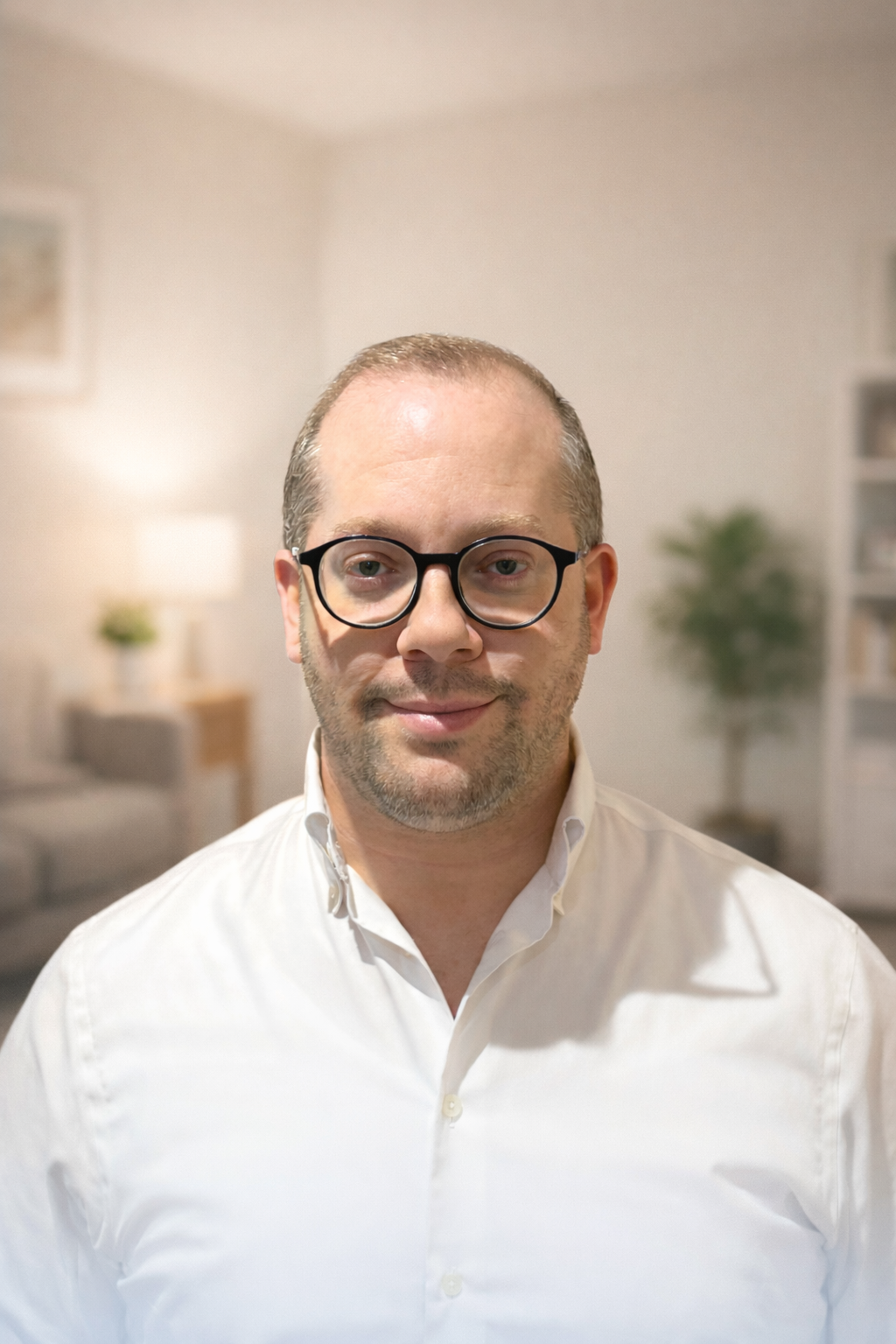 Portrait of a man with short hair, glasses, and a beard, wearing a white dress shirt, standing in a well-lit, cozy living room with blurred background decor. a resident doctor in our adhd, autism and general psychiatry team.