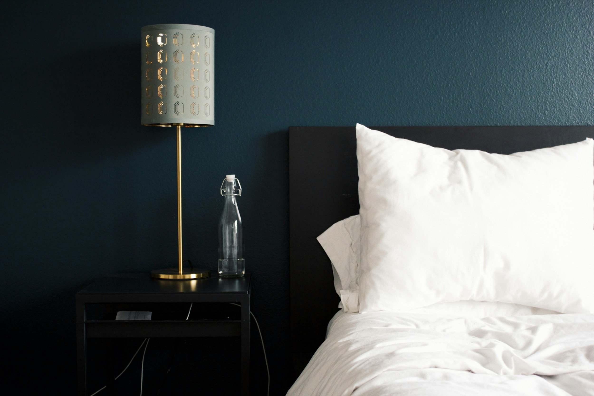 A bedside table with a gray nightstand, a modern table lamp with a perforated gray lampshade and a gold stand, a clear glass water bottle, and a white pillow and bed linens against a dark teal wall.