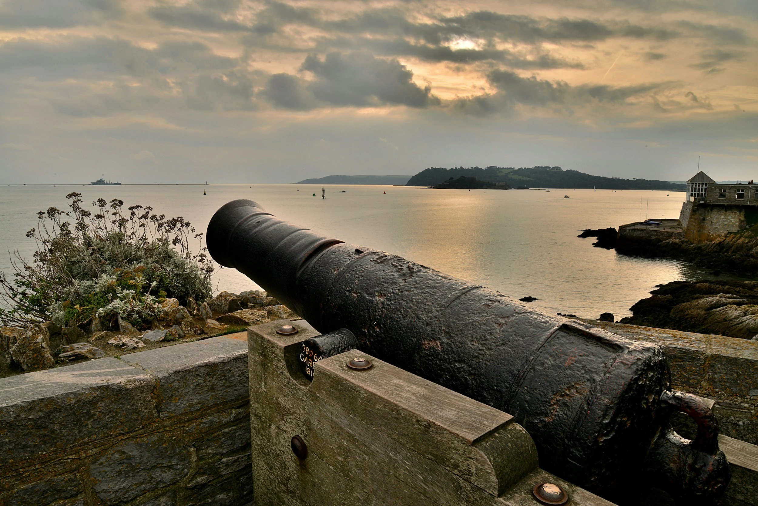 An old cannon points out towards the water on a stone platform near a seaside town. In the background, there is a body of water with ships, an island, and a cloudy sky at sunset.