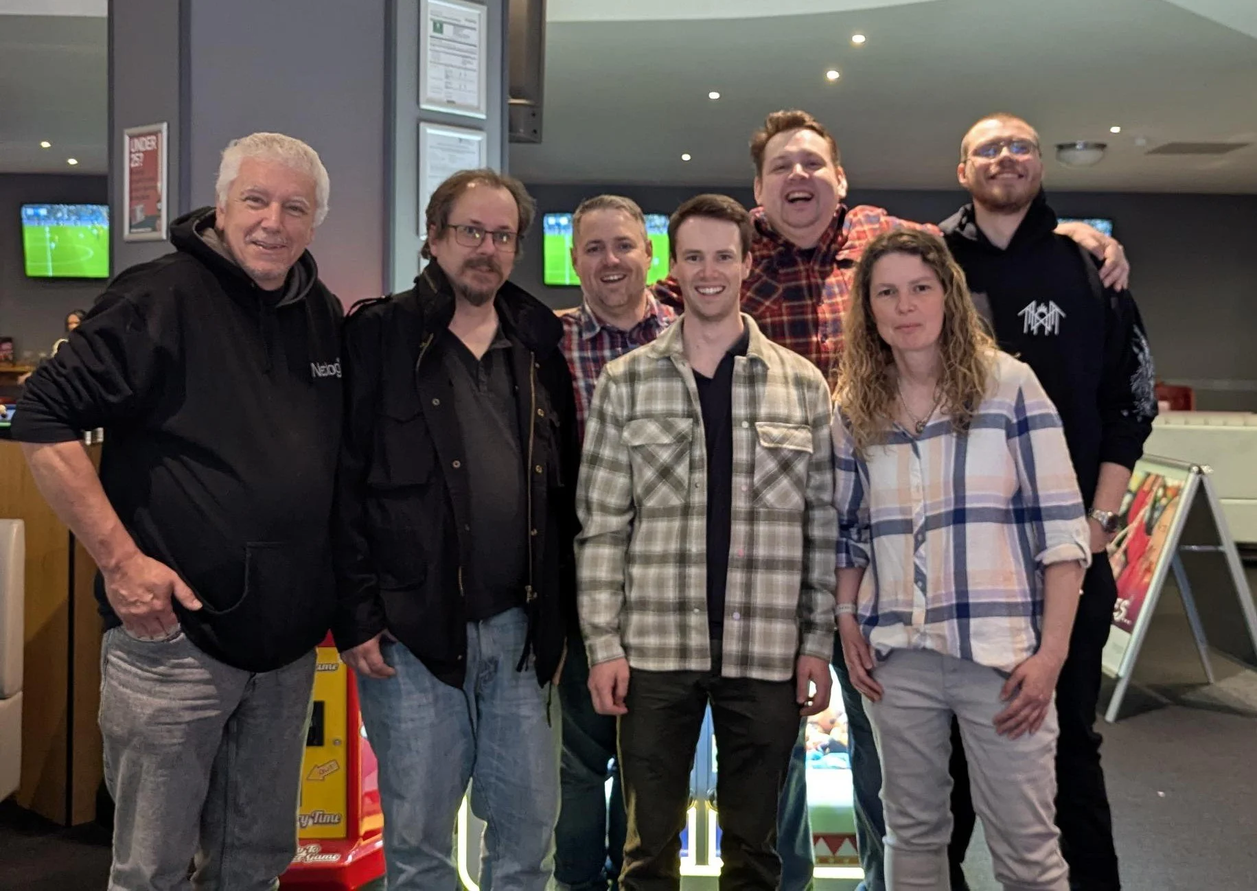 Group of seven people standing together, smiling for a photo in an indoor setting with TV screens showing sports in the background.