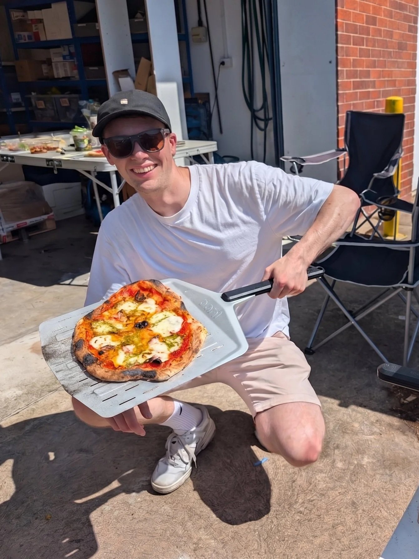 A man smiling and wearing sunglasses, a cap, a white t-shirt, and shorts, holding a pizza on a gray tray outdoors with a smile. There is a table with food in the background, and he is kneeling on the ground.