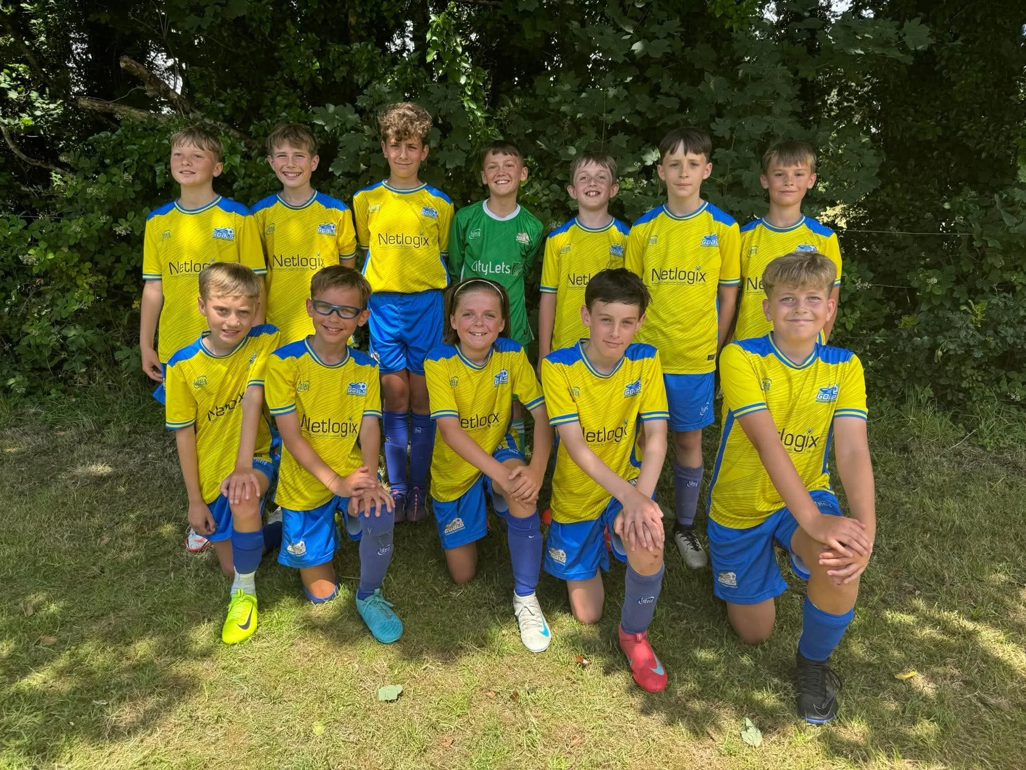 A group of young boys and girls in yellow and blue soccer uniforms posing outdoors in front of green trees.