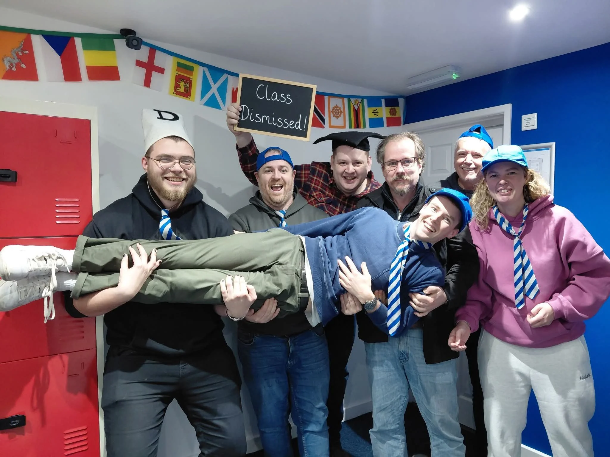 Group of six people posing inside a room with colorful national flags hanging on the wall, one person holding a blackboard sign that says "Class Dismissed!" The group appears to be celebrating or marking the end of a class or event.