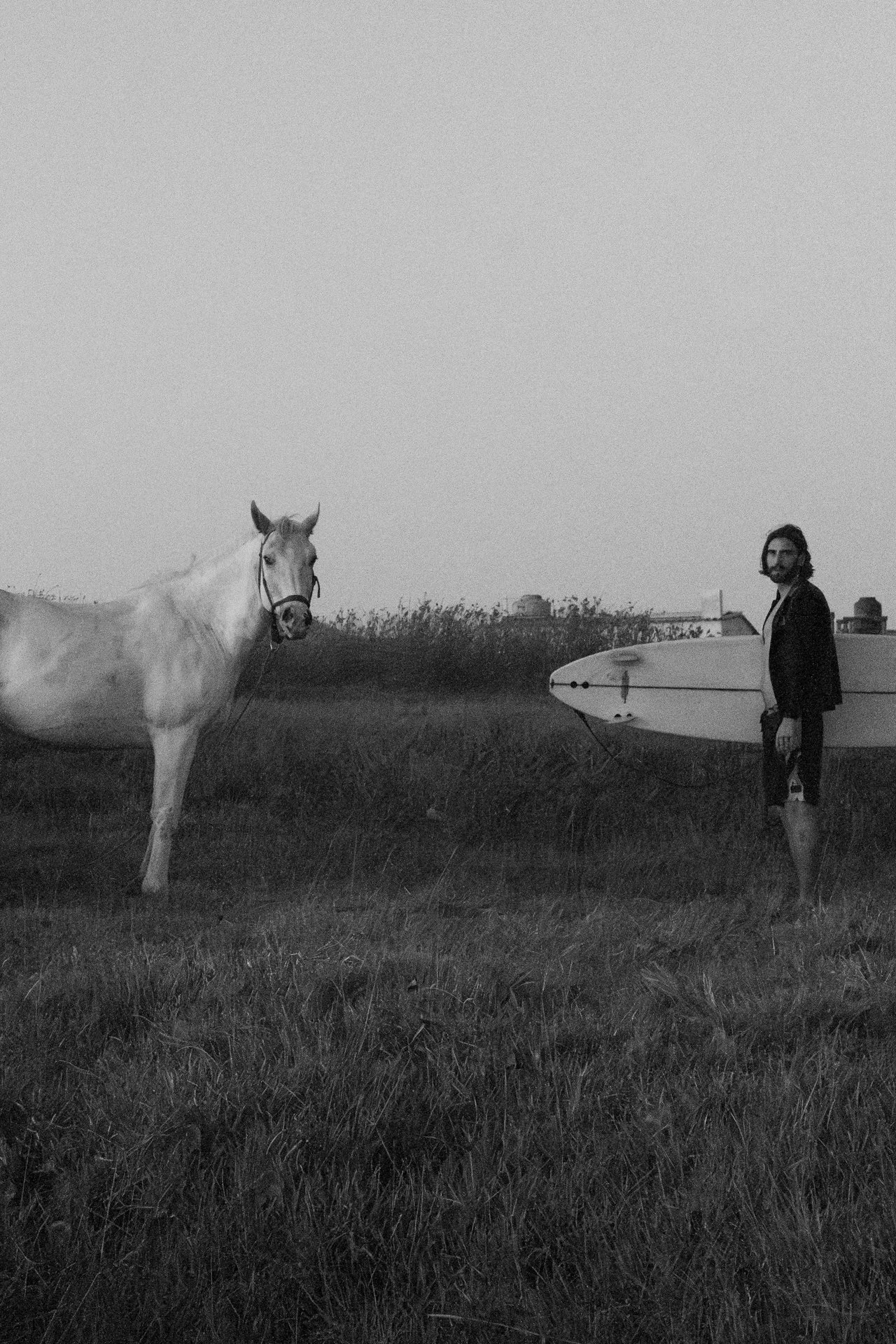 Black and white portrait of a surfer standing beside a horse in a rural landscape.