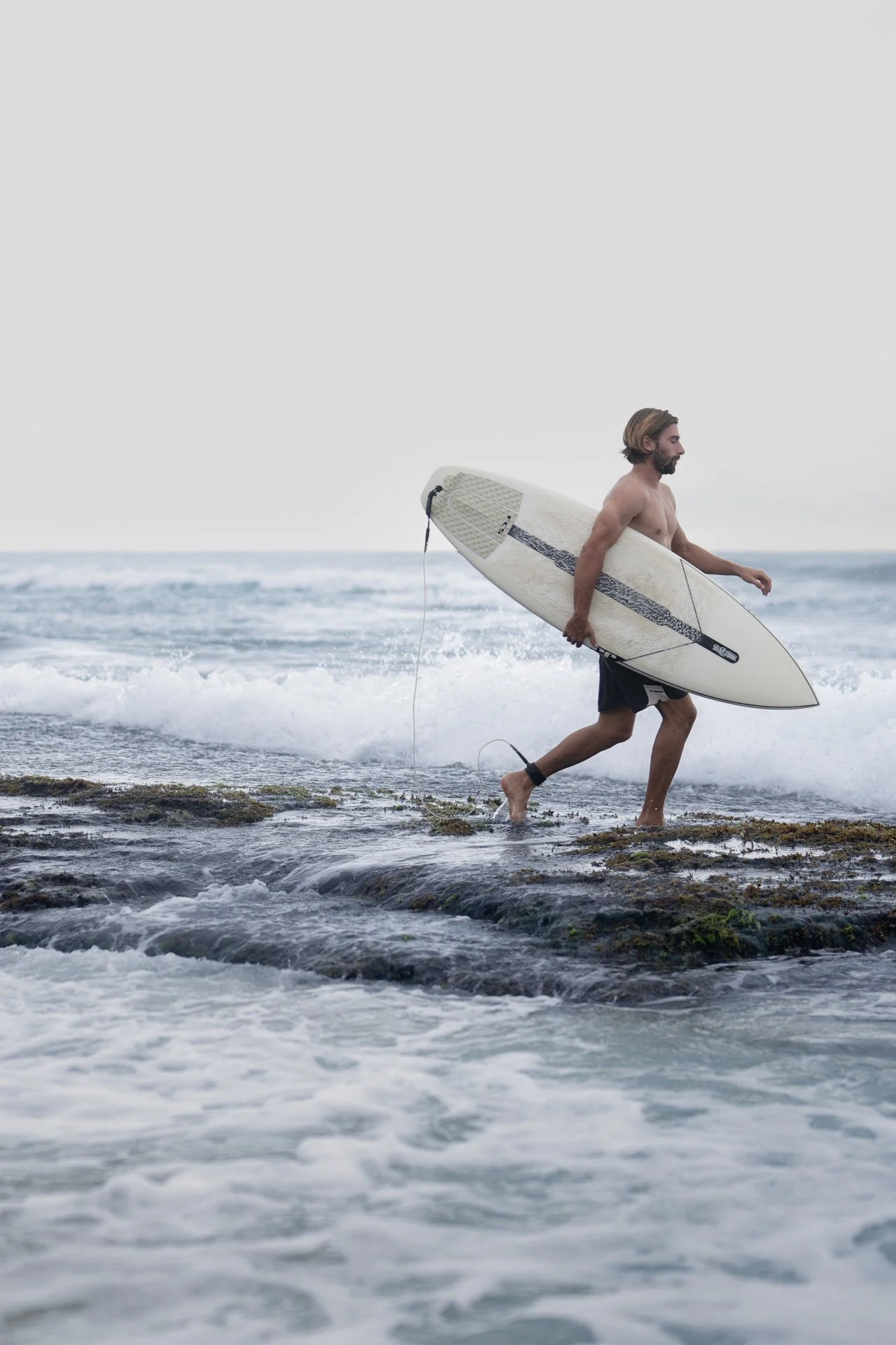 Surfer carrying a surfboard through shallow water along a remote shoreline.