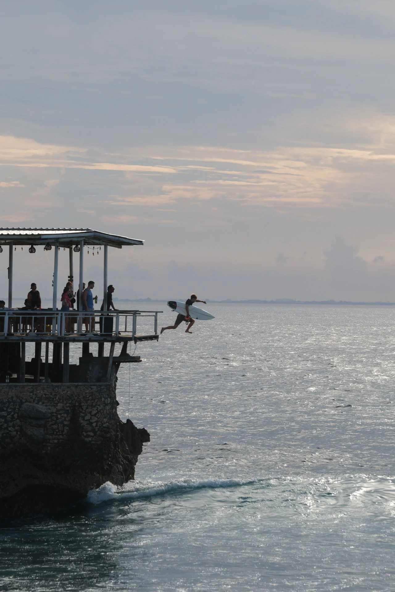 Surfer jumping from a coastal pier into the ocean at sunset.