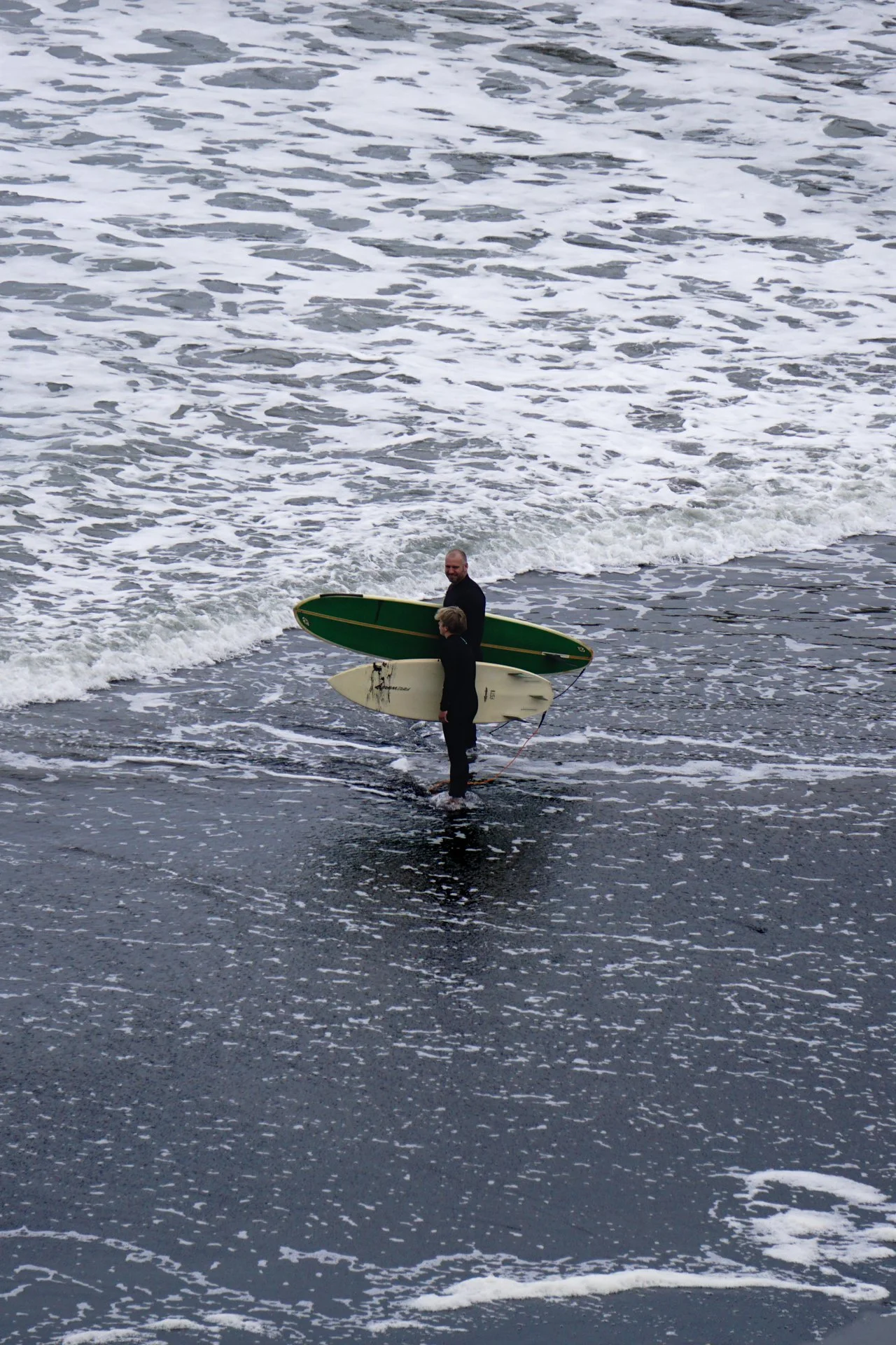 Two surfers walking into the ocean carrying surfboards, black sand beach, New Zealand. 