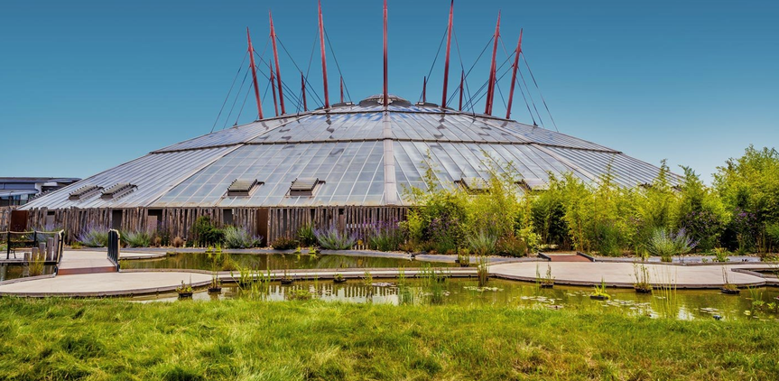 Un bâtiment rond avec une structure métallique, entouré de végétation et d'un petit plan d'eau, dans un paysage ensoleillé.