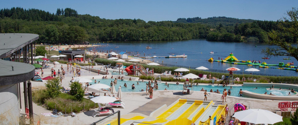 Piscine et espace aquatique au bord d'un lac, avec des gens nageant, jouant et profitant du soleil sous des parasols, en arrière-plan des bateaux et une aire de jeux aquatiques.