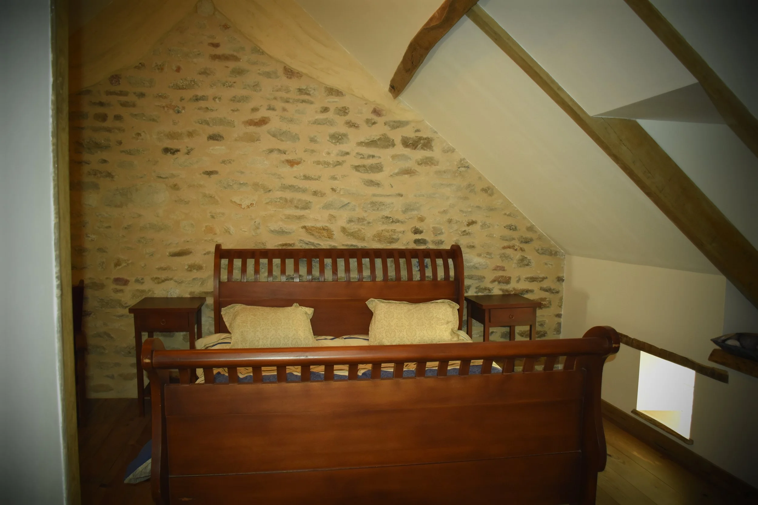 Chambre avec mur en pierre, lit en bois avec deux oreillers, deux tables de chevet, et une poutre en bois au plafond.