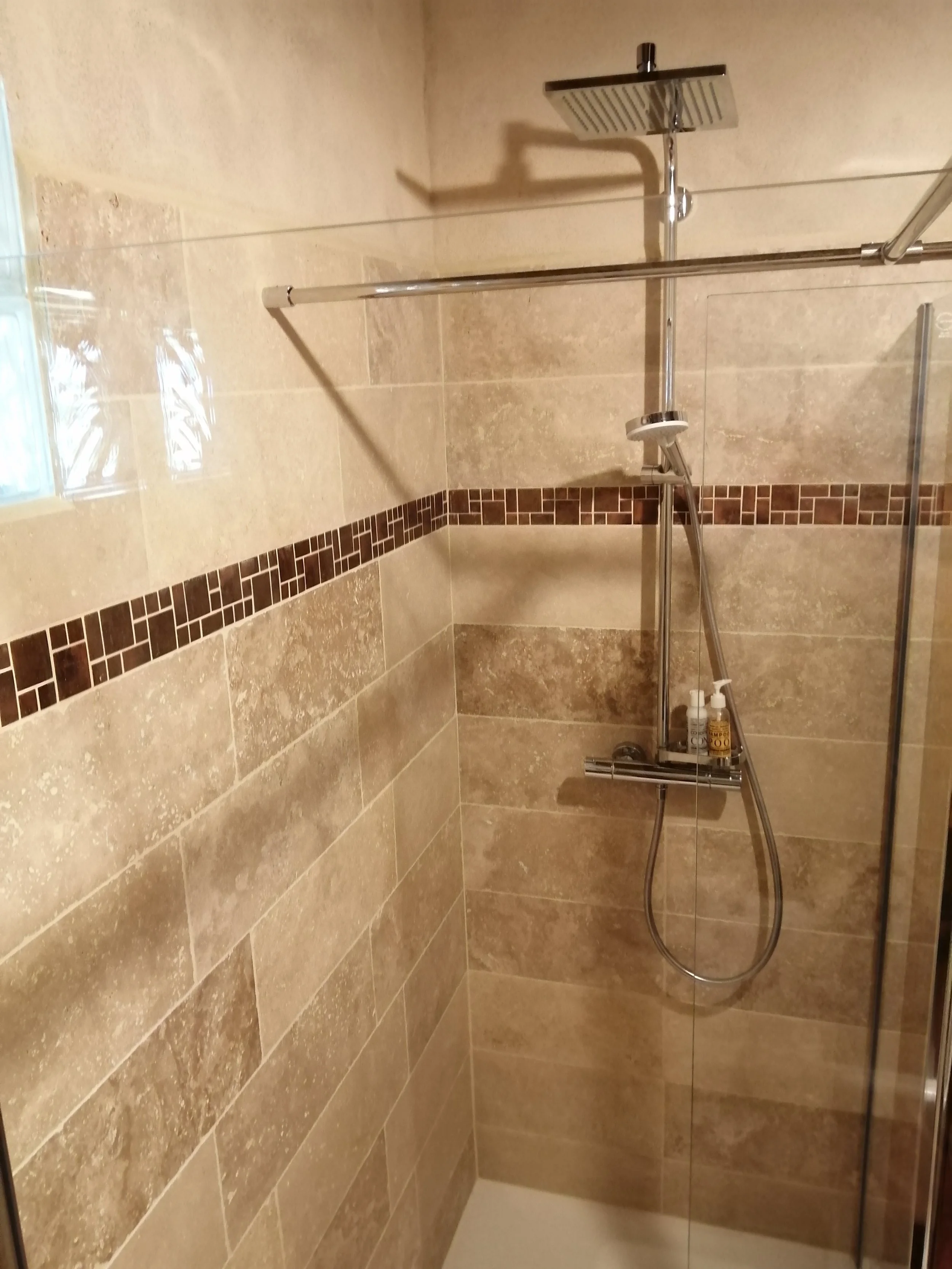 Douche avec paroi en verre, pomme de douche carré, murs et sol en carreaux beige et marron.