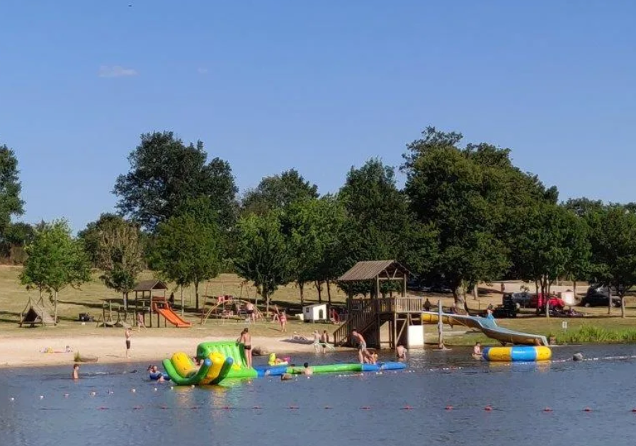 Les enfants jouent dans une zone de baignade avec des structures gonflables dans un lac ou une piscine, entourée d'arbres et d'espaces verts, par une journée ensoleillée.
