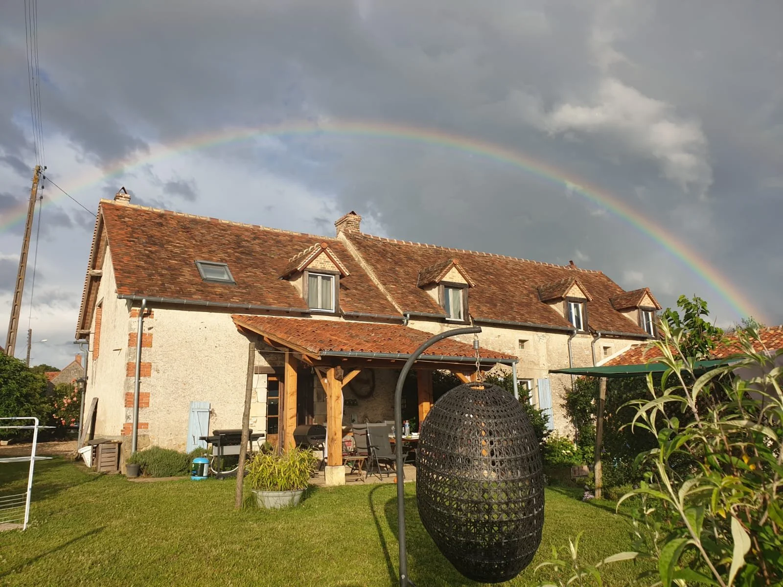 Maison traditionnelle en pierre avec un toit en tuiles rouges, sous un ciel nuageux avec un arc-en-ciel visible en arrière-plan.