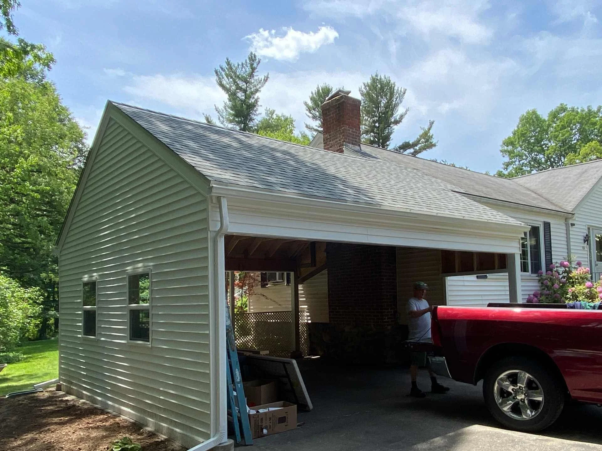 A house with white vinyl siding and a shingled roof, with an open garage area completed after the existing structure was destroyed by a falling tree in Holden, MA. Green trees and a partly cloudy sky are in the background.
