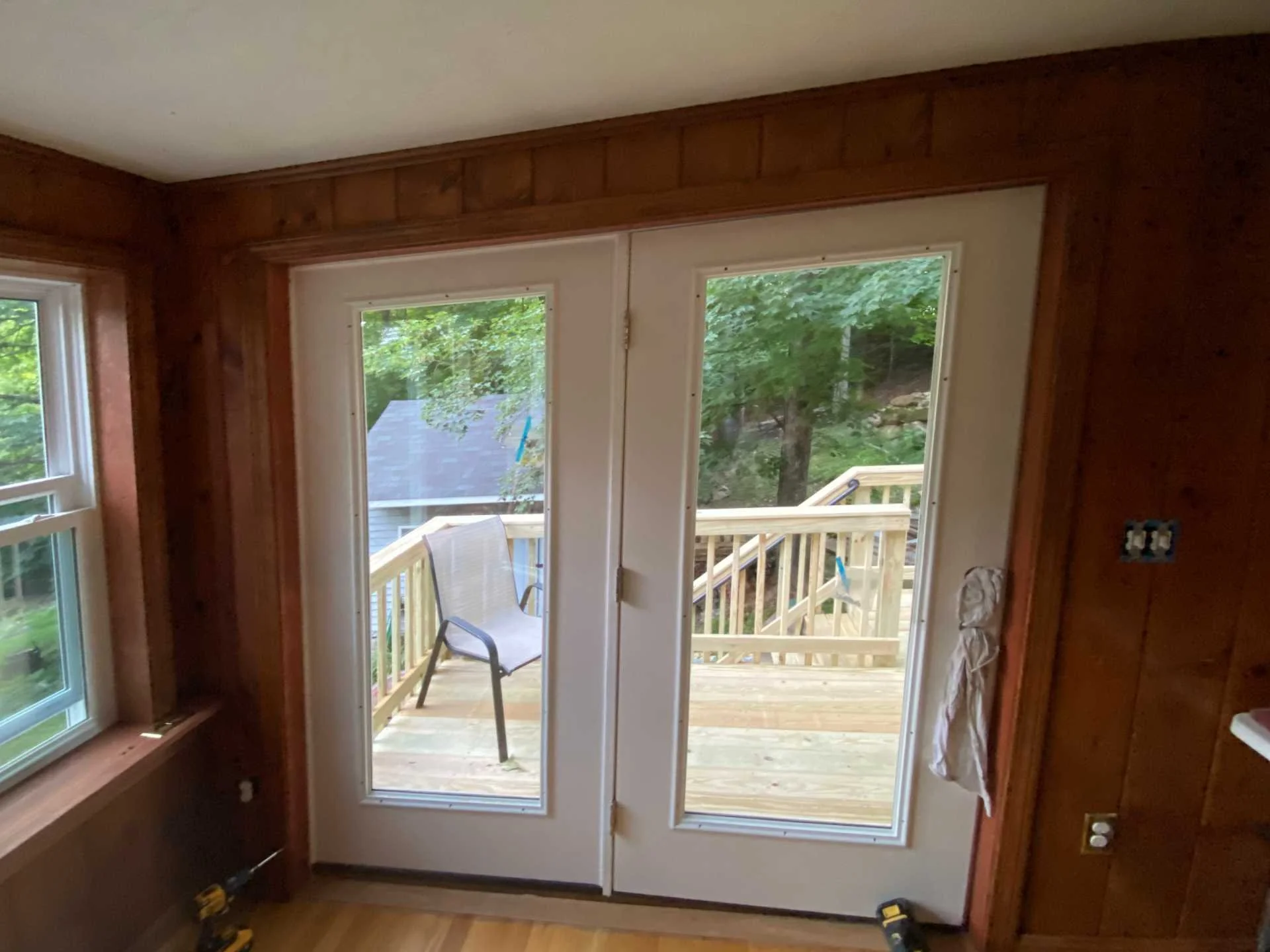 A set of glass patio doors with a center hinge leading to a wooden outdoor deck with steps, surrounded by green trees, inside a room with wooden walls and two new windows in Sterling, MA