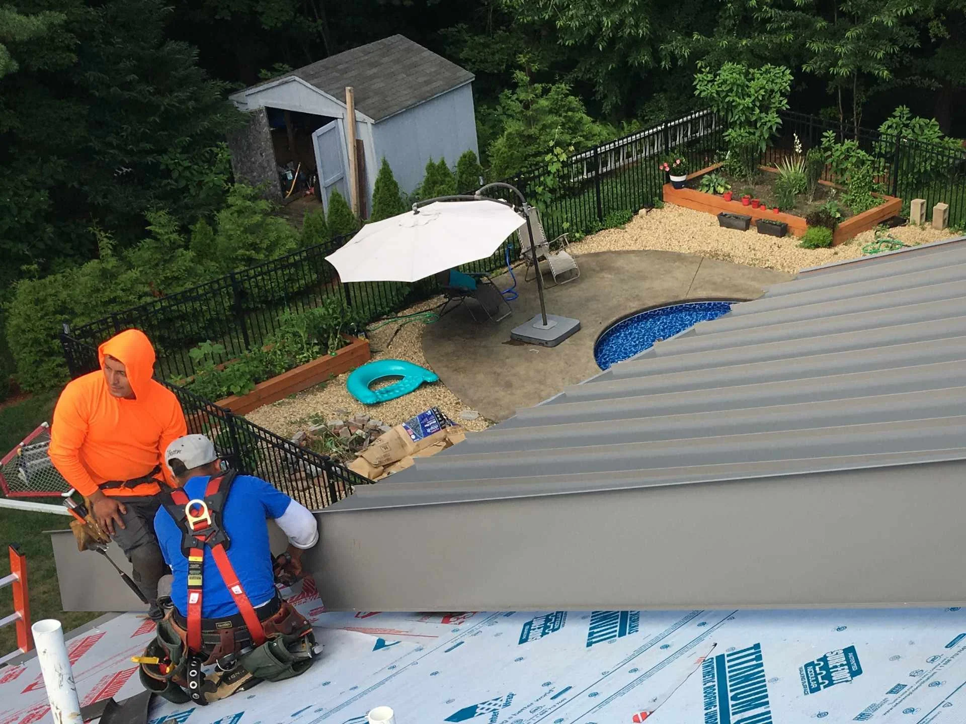 Construction workers install standing-seam metal roofing materials on a house, with a backyard garden, a small pool, and a shed visible in the yard in Fitchburg, MA