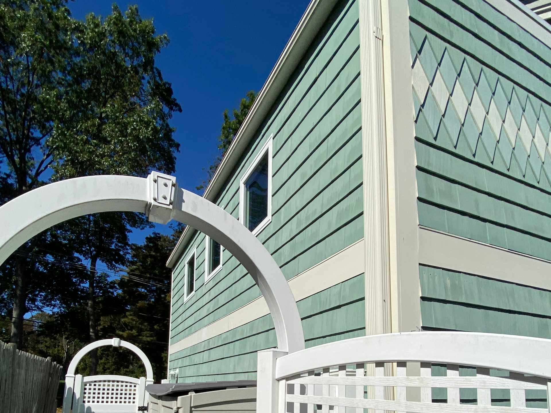 View of a light green house green cedar shake siding and PVC trim in Belmont, MA with a white fence and decorative arch in the foreground. There are trees in the background with a clear blue sky.