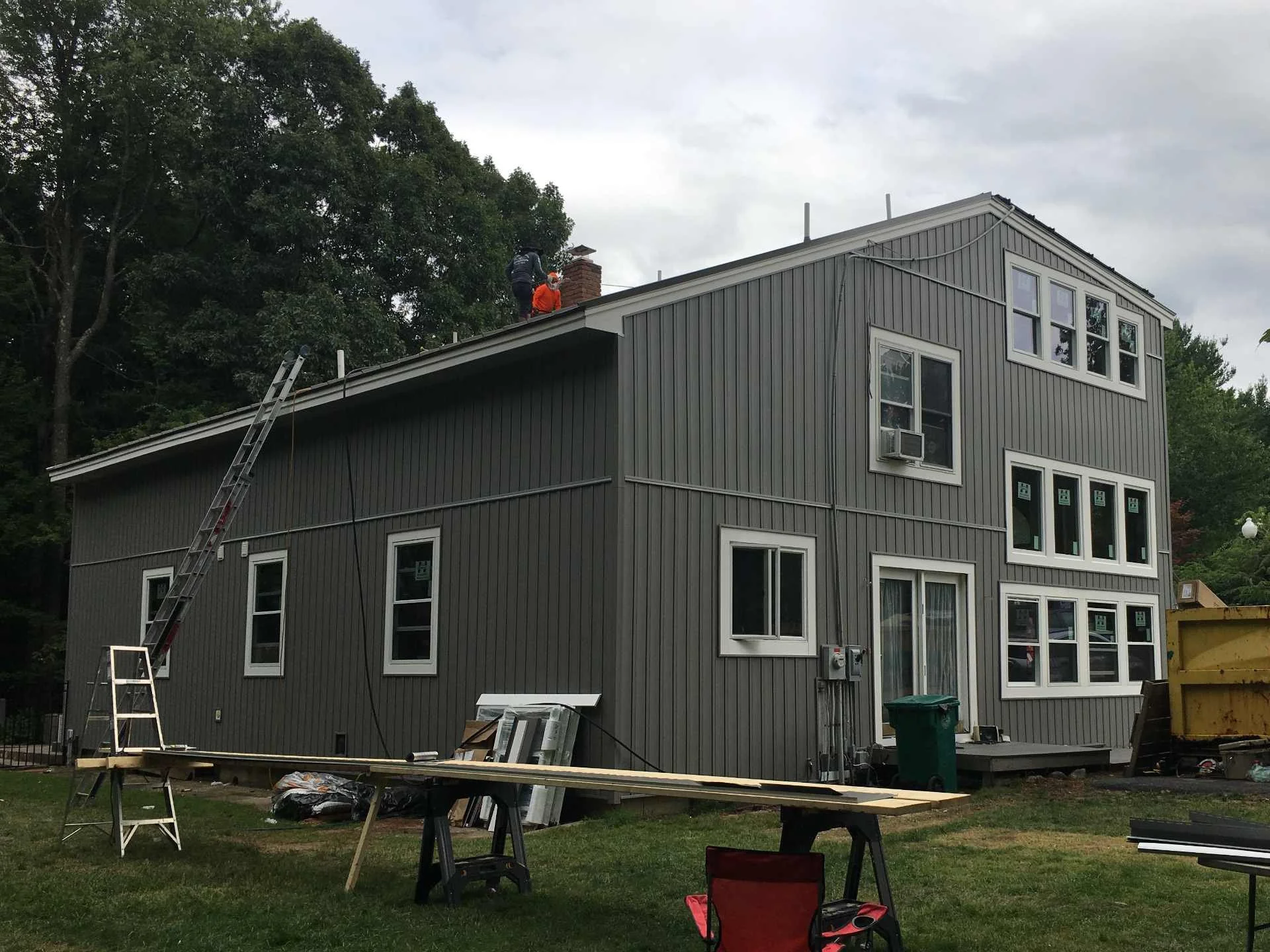 Construction workers finishing the standing-seam metal roof of a gray, multi-story house in Fitchburg, MA after completing the install of large windows and vertical board and batten vinyl siding. Image is during daytime; construction tools, a ladder,
