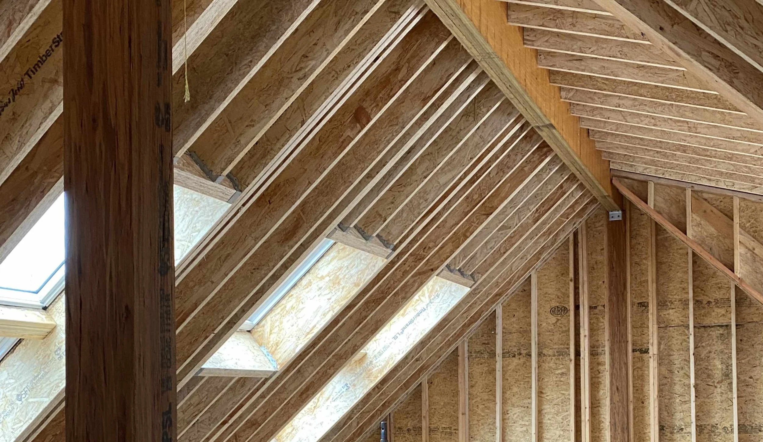 Interior of a building under construction with wooden roof framing, visible roof trusses, and a skylight window.