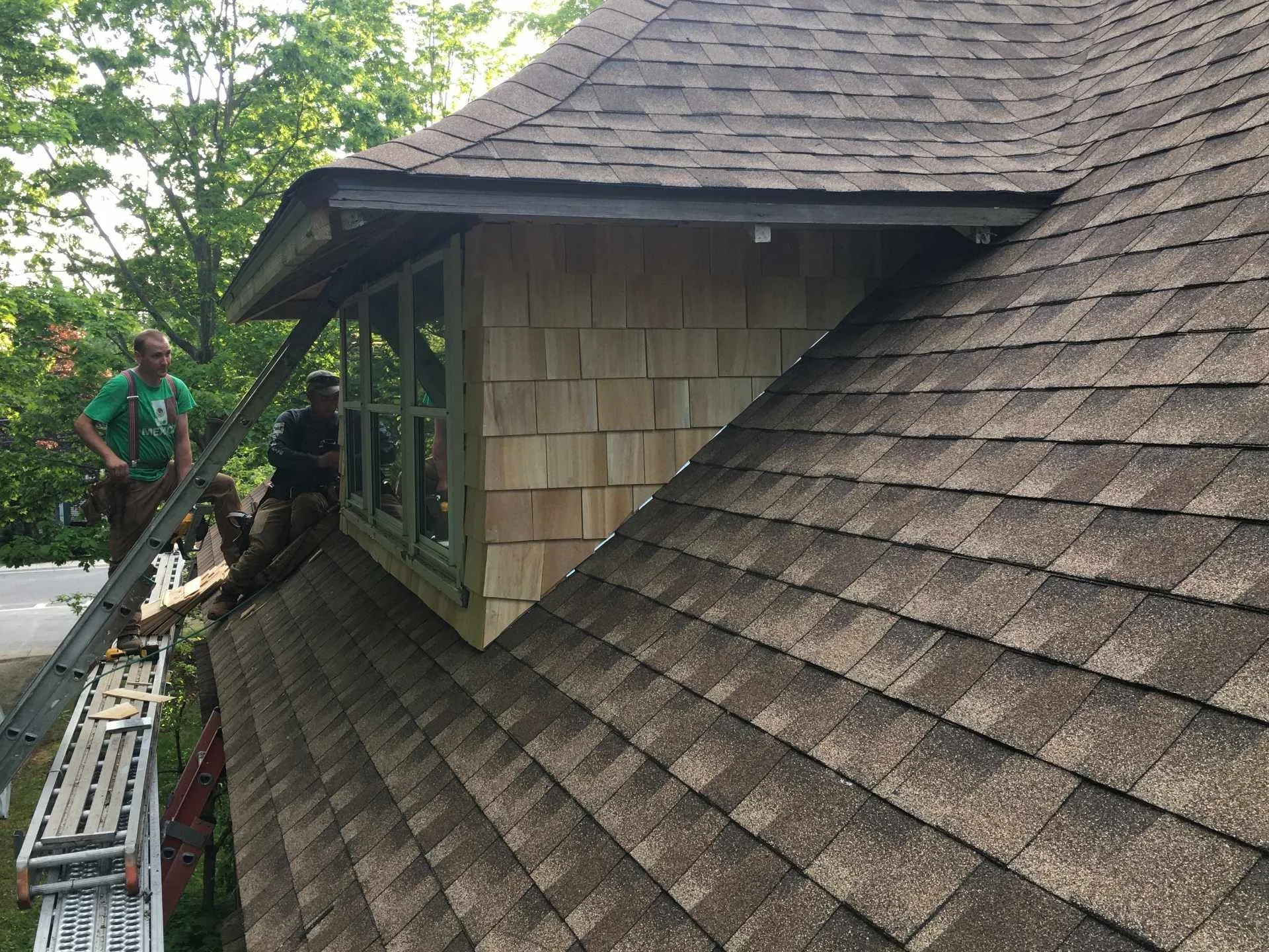 Construction workers installing a new attic window in a residential house, with roofing shingles visible on the roof.