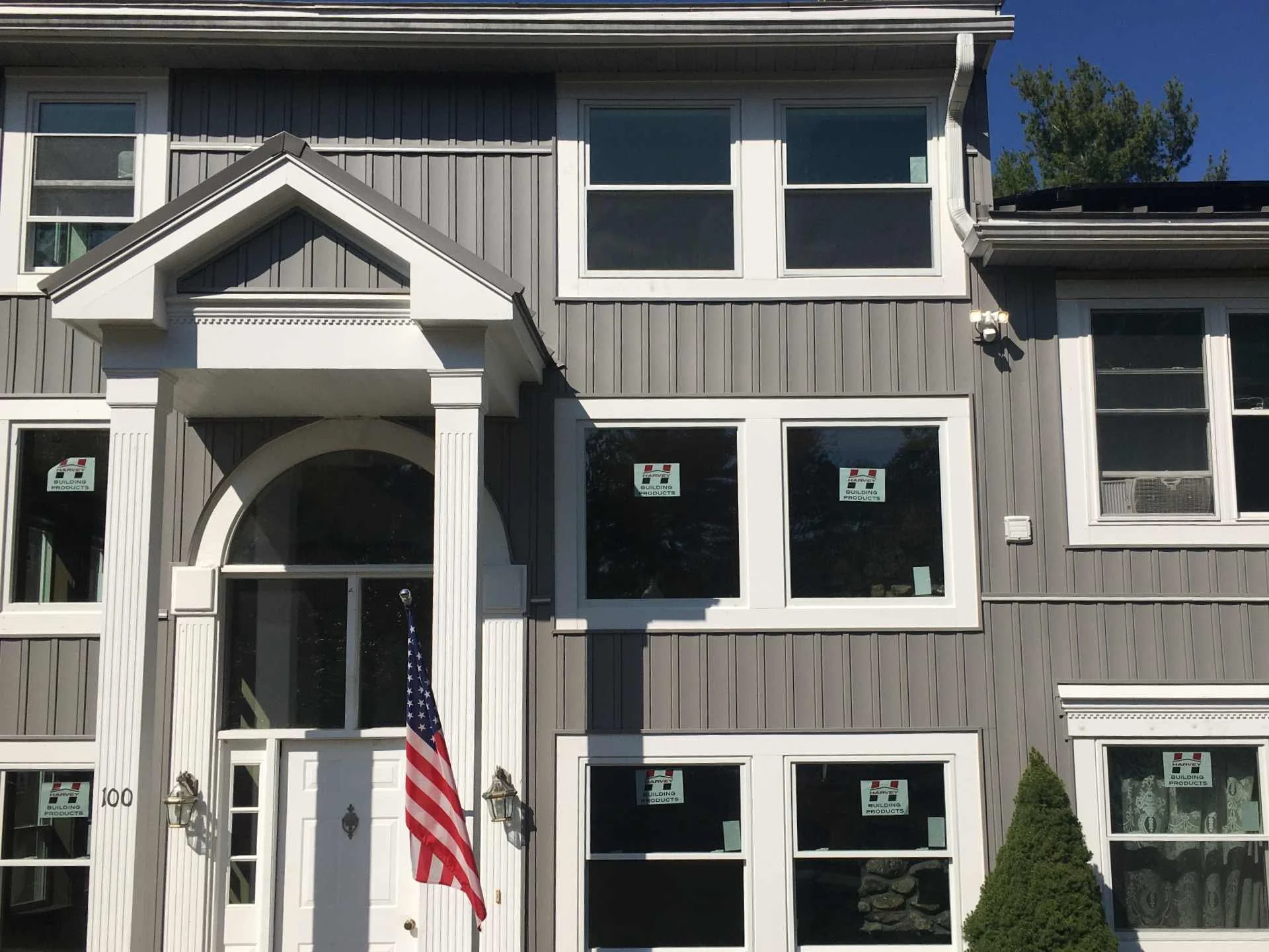 Front view of a gray multi-story house with white trim, an arch above the entrance, an American flag on a pole near the door, and several windows with signs that read 'Harvey Building Products' in Fitchburg, MA installed by McGrath Construction