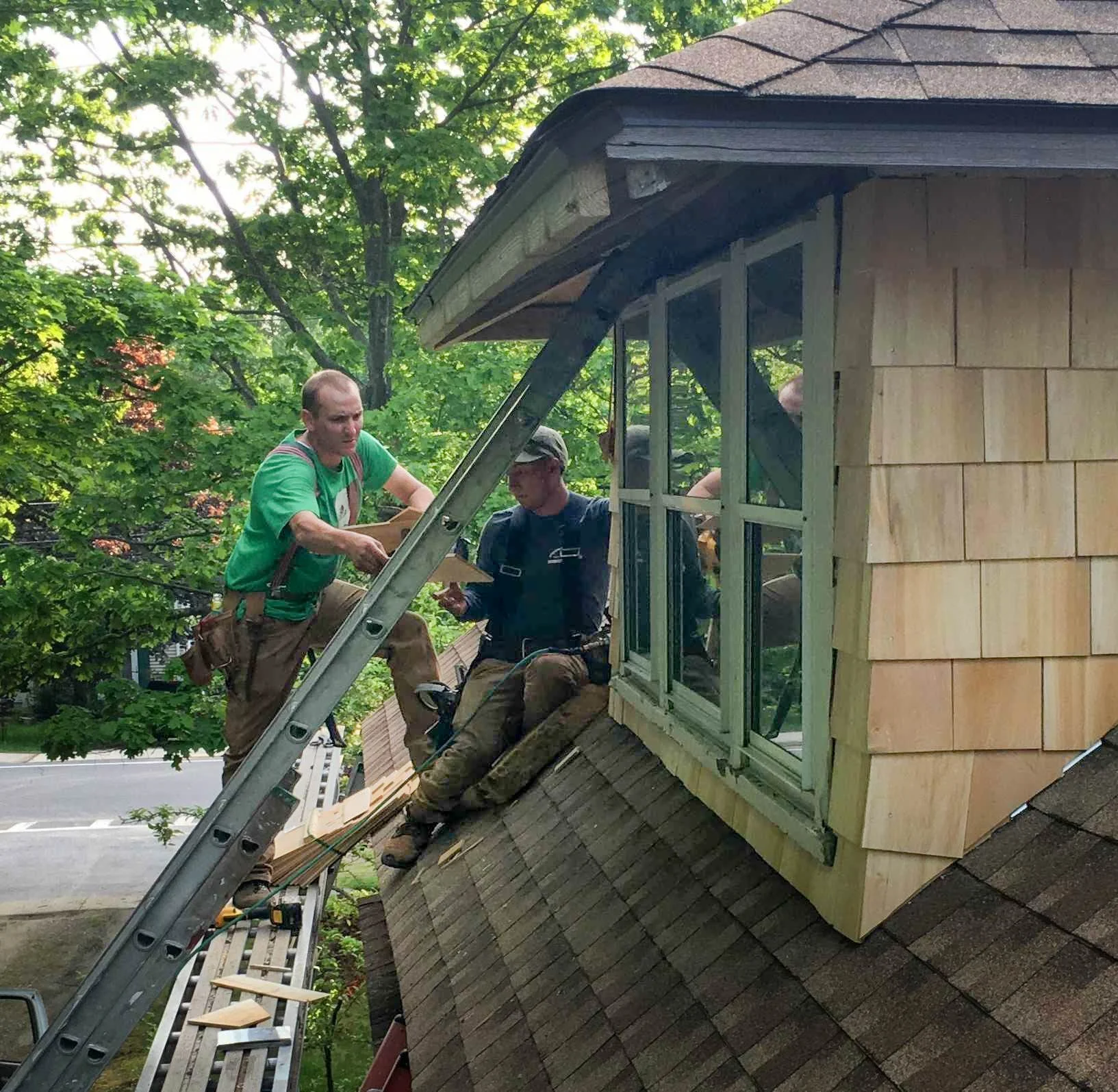 Two men working on the roof of a house during daytime, with green trees in the background. They are finishing the cedar shake siding on the dormer after they had completed the roof replacement due to storm damage
