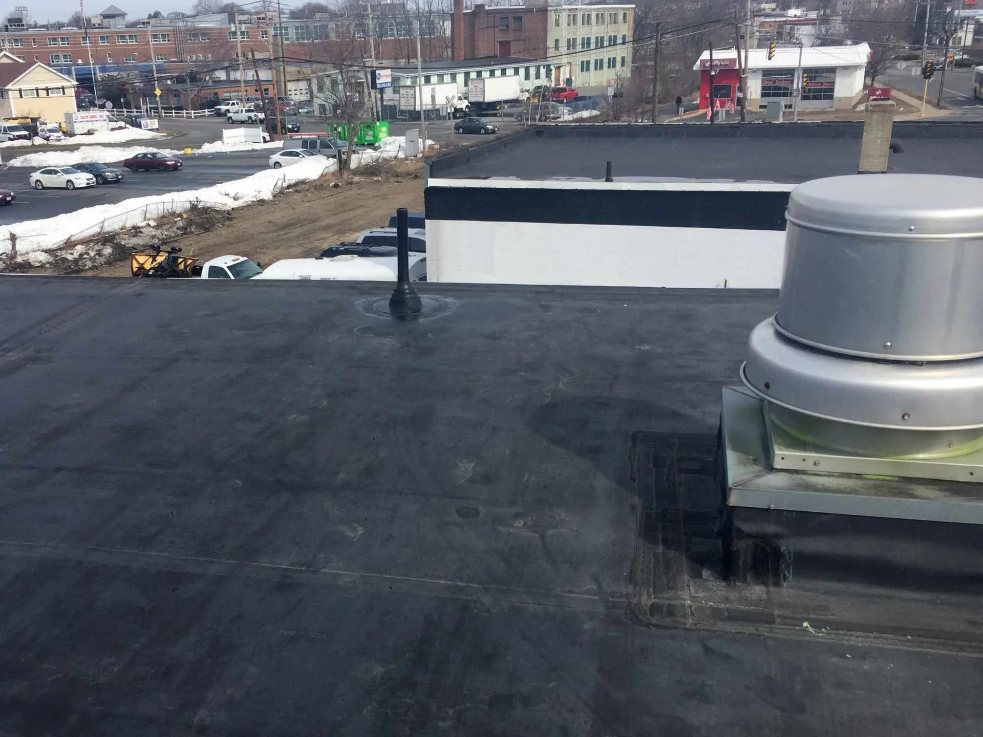 A new EPDM rubber roof with a metal vent in the foreground in Dracut, MA with a parking lot with several parked cars, and a city street with moving cars, buildings, and snow patches on the ground.