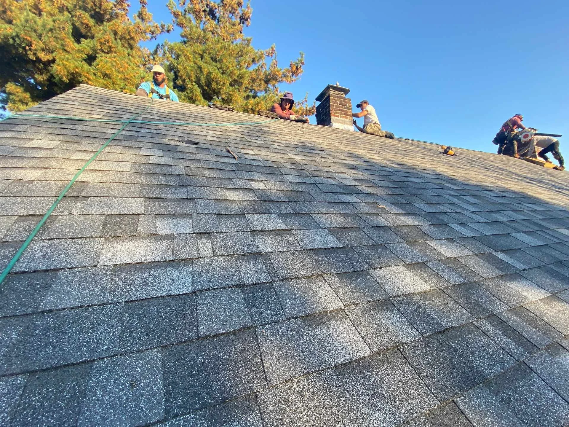 Four workers finishing a sloped asphalt roof replacement and installing new lead flashing on a chimney in Belmont, MA during daytime, with a bright blue sky and large trees in the background.