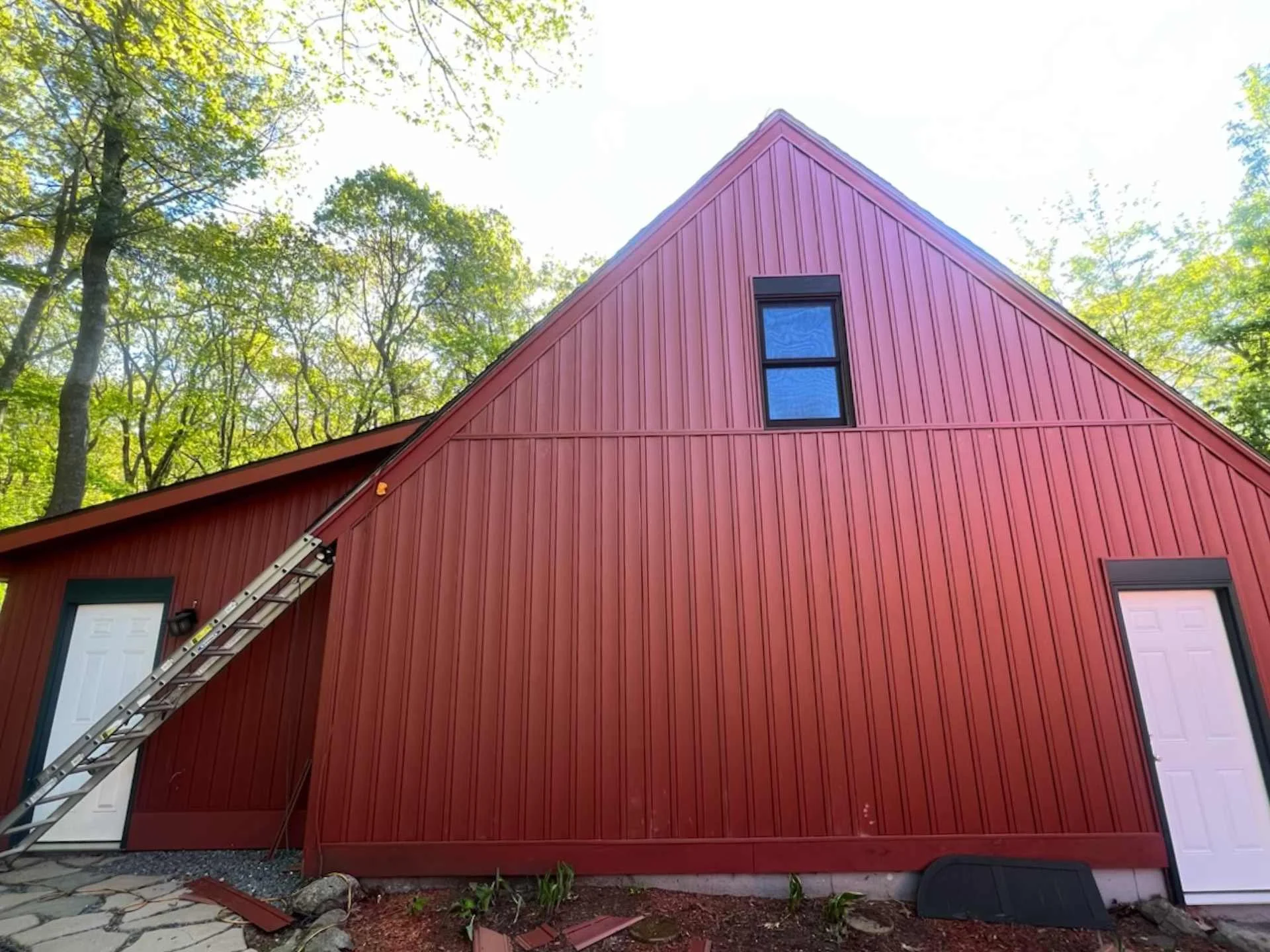 Red garage with vertical siding, two visible white garage doors, a ladder leaning against the house, and trees with green leaves surrounding it on Wachusett Mountain in Princeton, MA