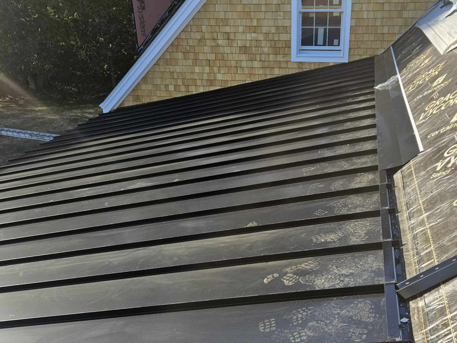Close-up of a black standing-seam metal roof install and cedar shake siding install with a white-framed window in Westford, MA