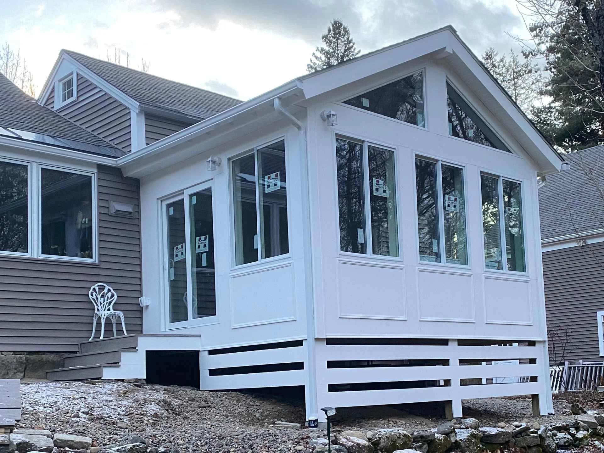 A house with a white four-season sunroom addition, multiple large windows, a gable roof, and a small porch with a white chair and steps, surrounded by a rocky and partially snow-covered ground with trees in the background in Winchendon, MA
