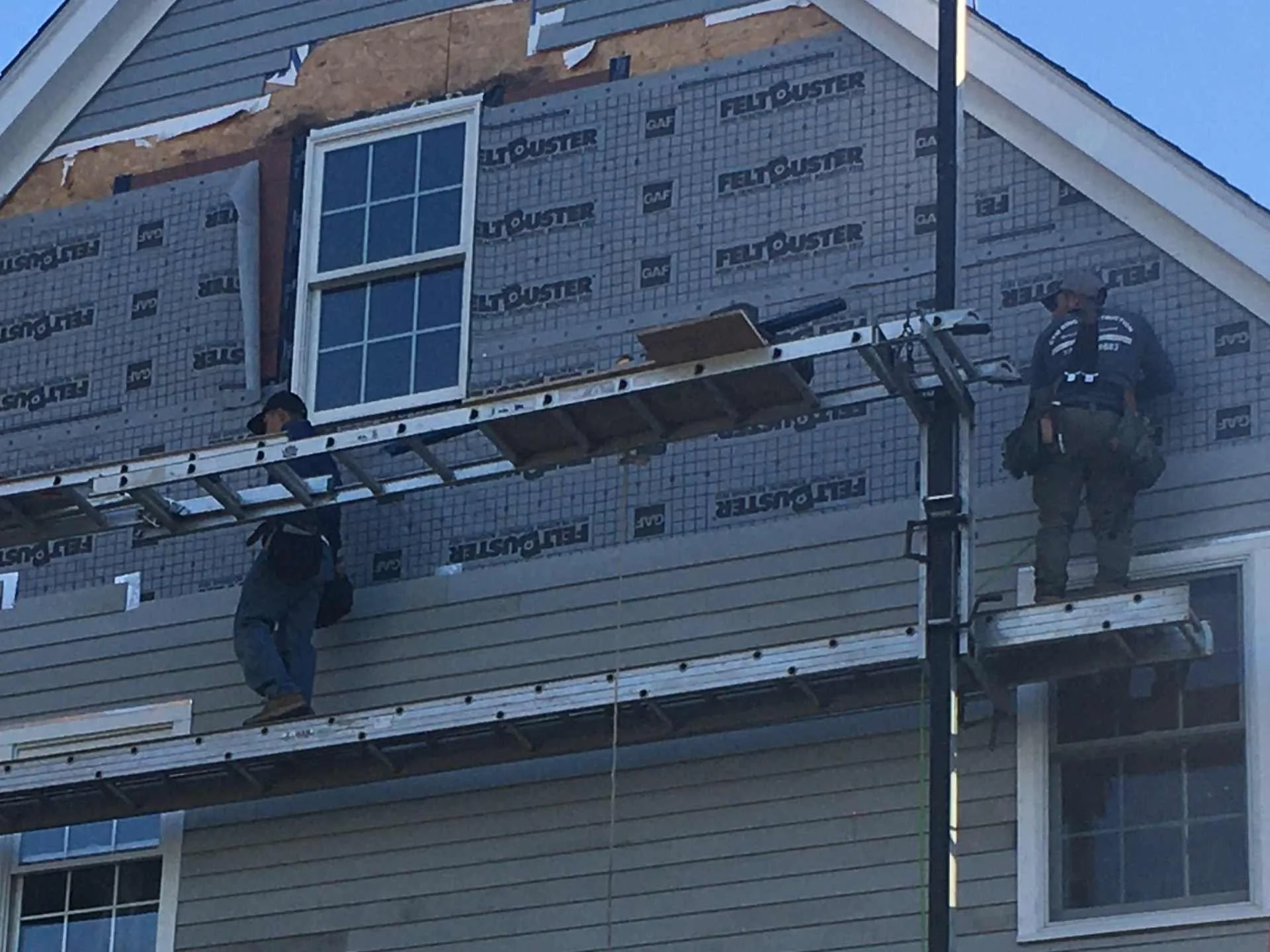Construction workers on scaffolding installing siding on a building with a window, with the building covered in weatherproofing material labeled 'FeltBuster'.