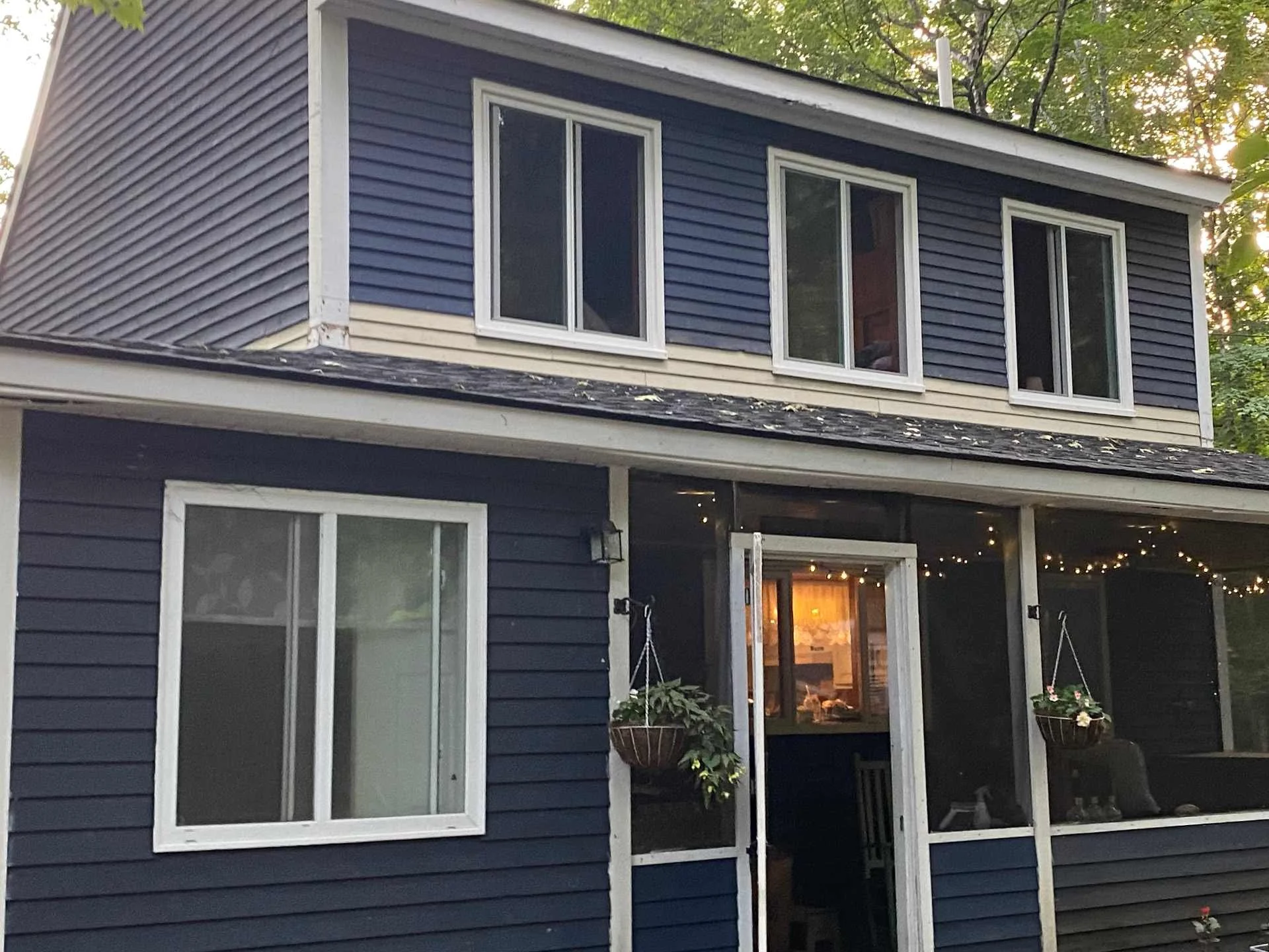 A two-story house with blue siding and white trim, featuring large sliding glass doors on the ground floor and three windows on the upper floor. There are hanging flower baskets on the porch and trees in the background.
