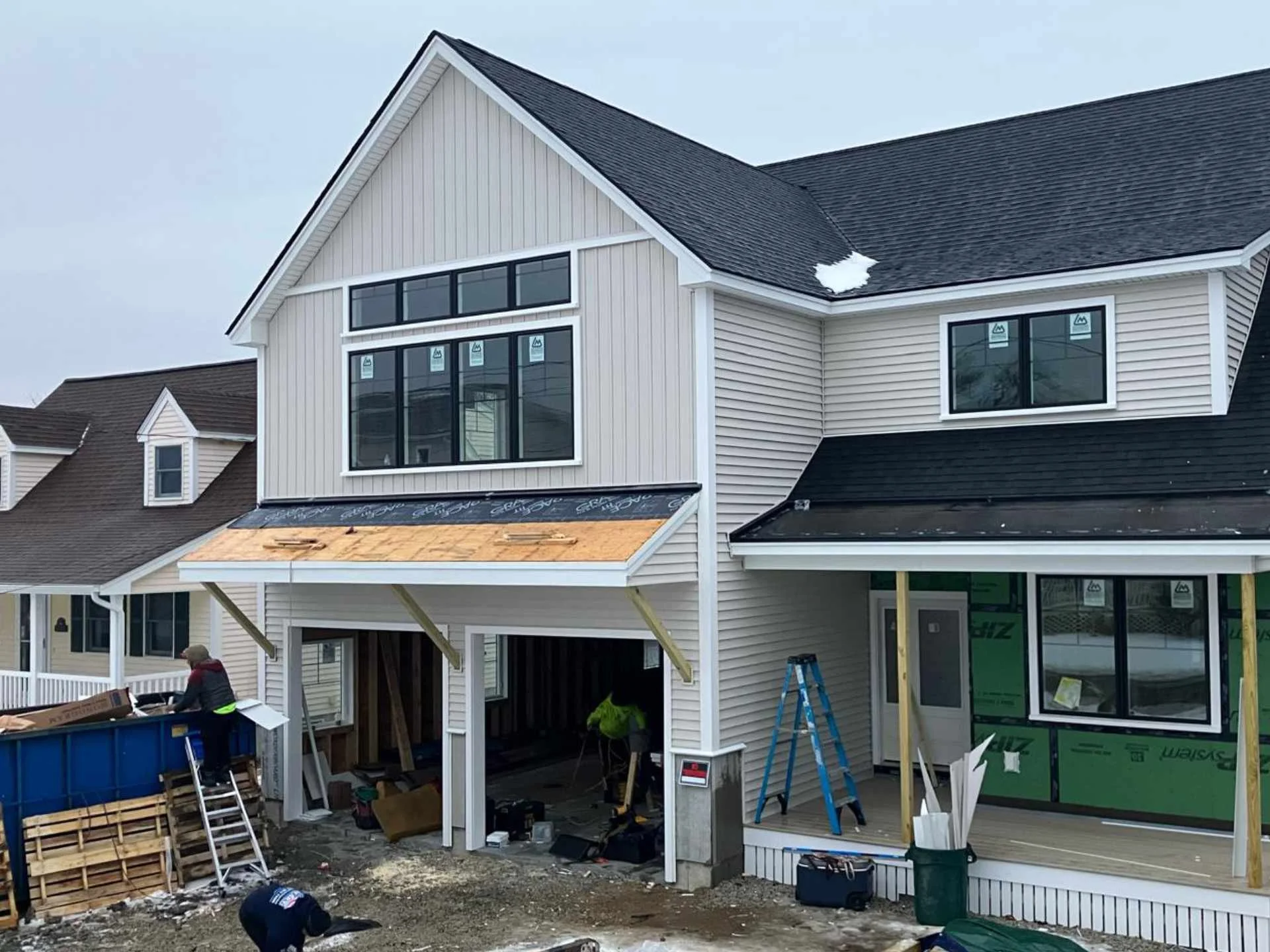 Construction workers working on the exterior of a multi-story house in Westford, MA with beige siding, black roofing, and large windows, in a residential area.