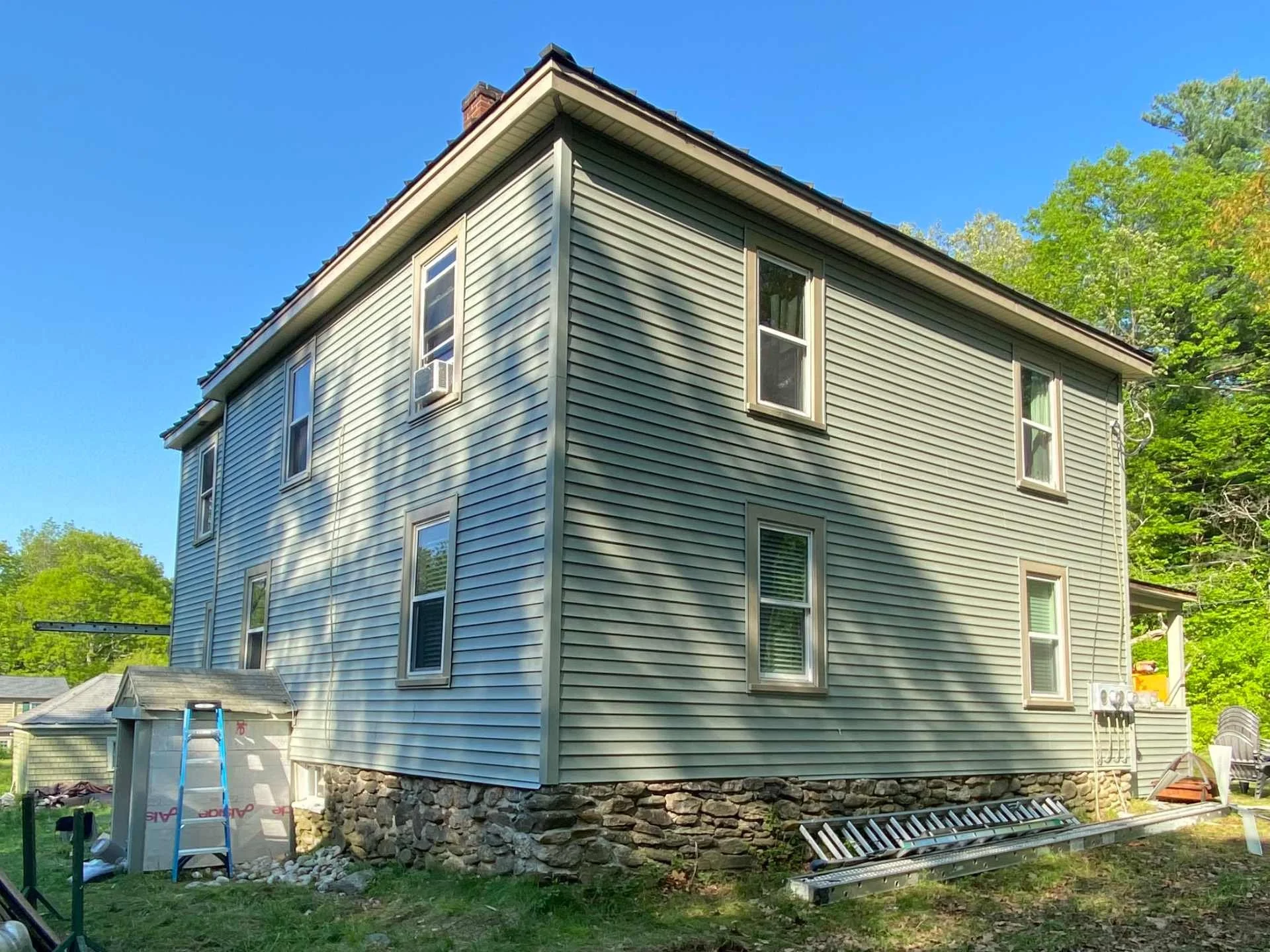 Three-story house in Princeton, MA with newly installed seagrass siding and white trim, surrounded by green trees, under a clear blue sky.