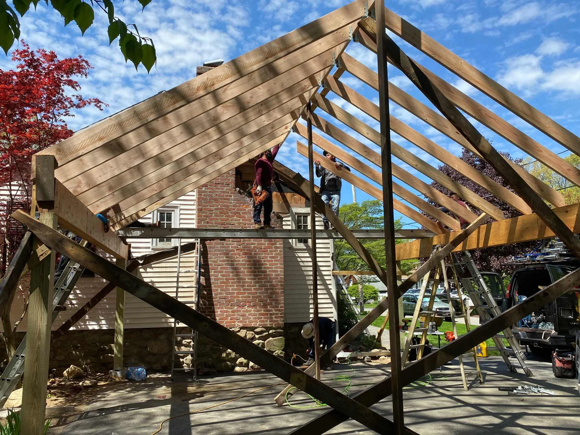 Construction workers building the roof of a carport that was destroyed by a falling tree in a storm in Holden, MA The roof's wooden frame is partially completed, with blue sky and clouds above. Tools and scaffolding are visible around the site.