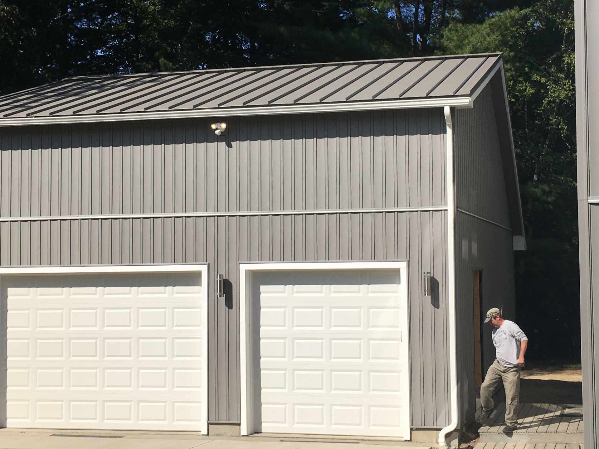 A brand new modern gray garage in Fitchburg, MA completed by McGrath Construction with two white garage doors, a standing-seam metal roof, Certainteed board and batten vinyl siding, and a security light on the wall. The business owner with a hat walk