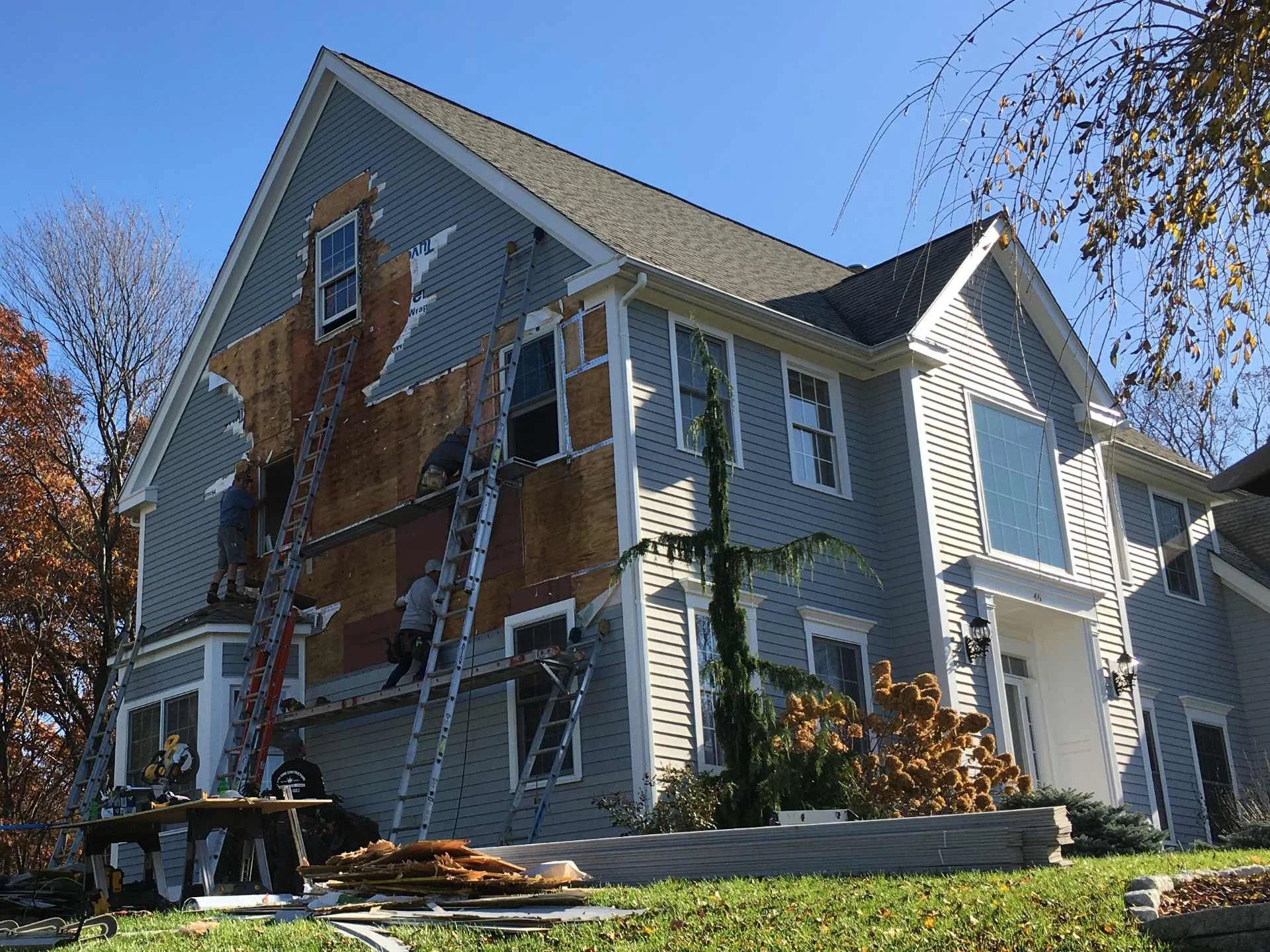 Workers replacing or repairing the exterior siding and insulation of a multi-story house. Ladder and construction tools are visible, and part of the house's siding is removed, exposing the underlying structure.