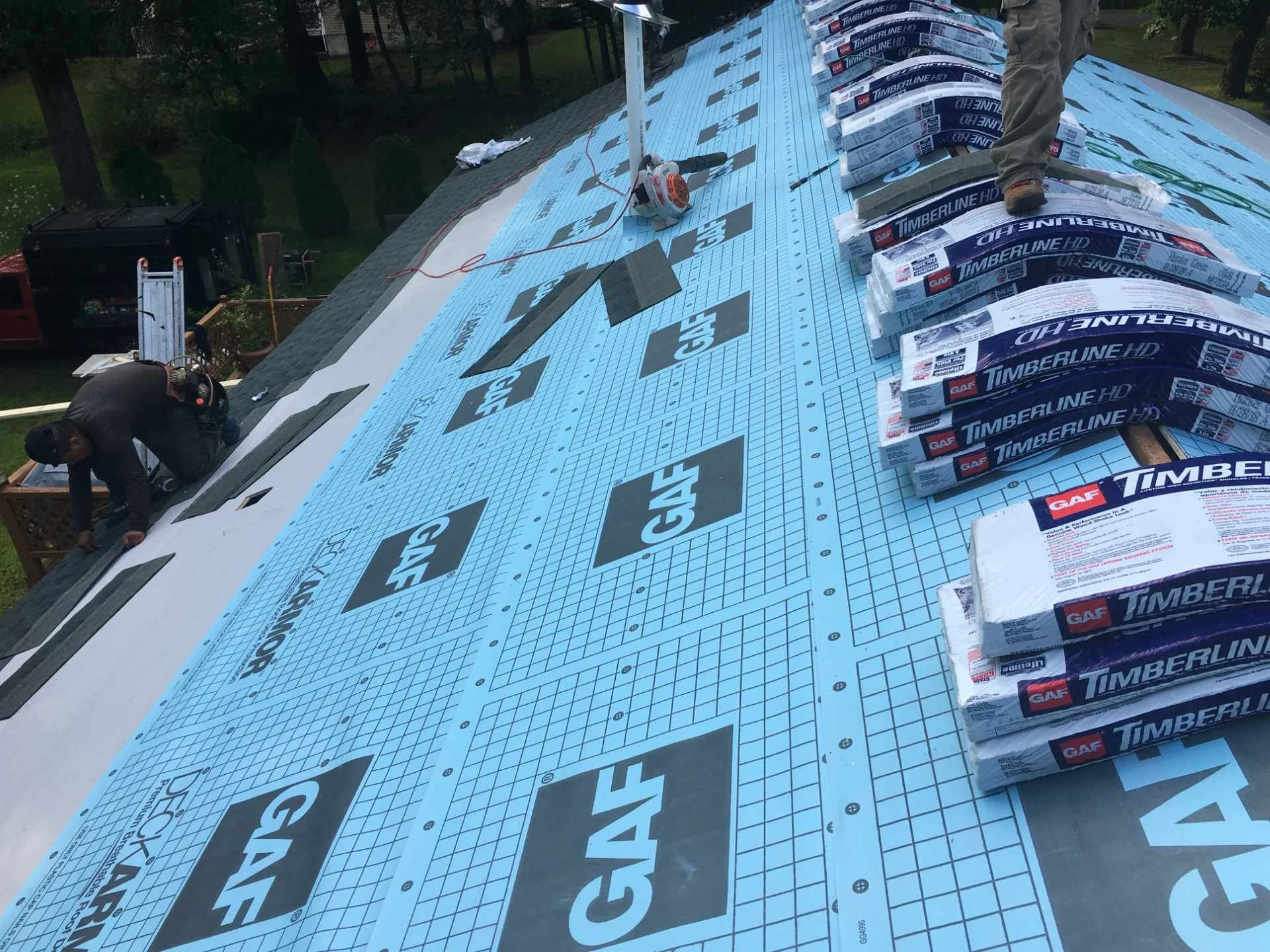 Construction workers installing roof shingles on a house roof in Tewksbury, MA with bundles of Timberline HD shingles and roofing tools.
