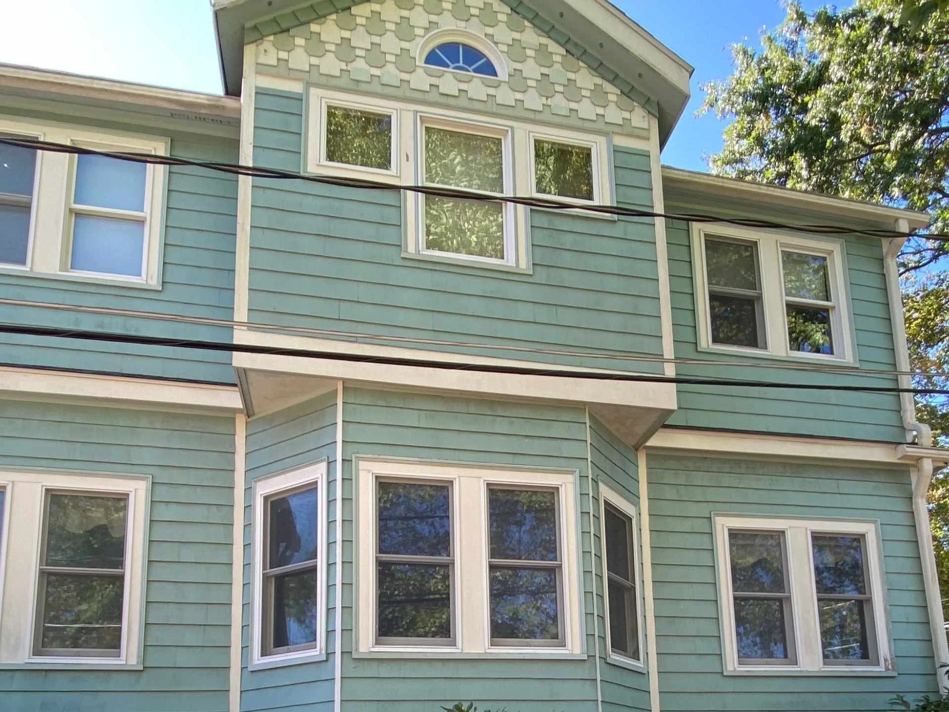 A close-up of a multi-story house with teal siding and white window frames. The house features several windows, including a bay window and a rounded window above. A tree with green leaves is visible on the right side, and blue sky is in the backgroun