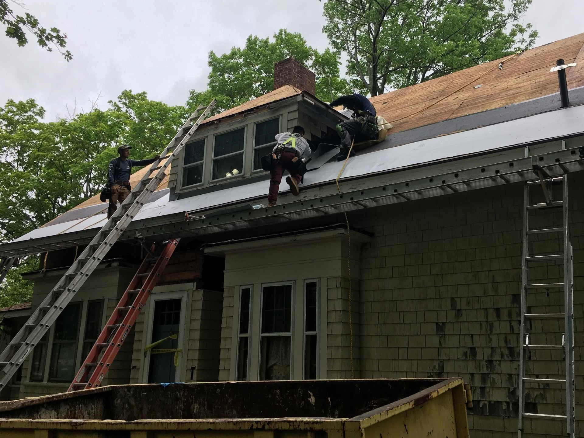Three construction workers on a residential roof under renovation, with one worker on a ladder and two working on the roof near a chimney, surrounded by trees and construction equipment in Templeton, MA