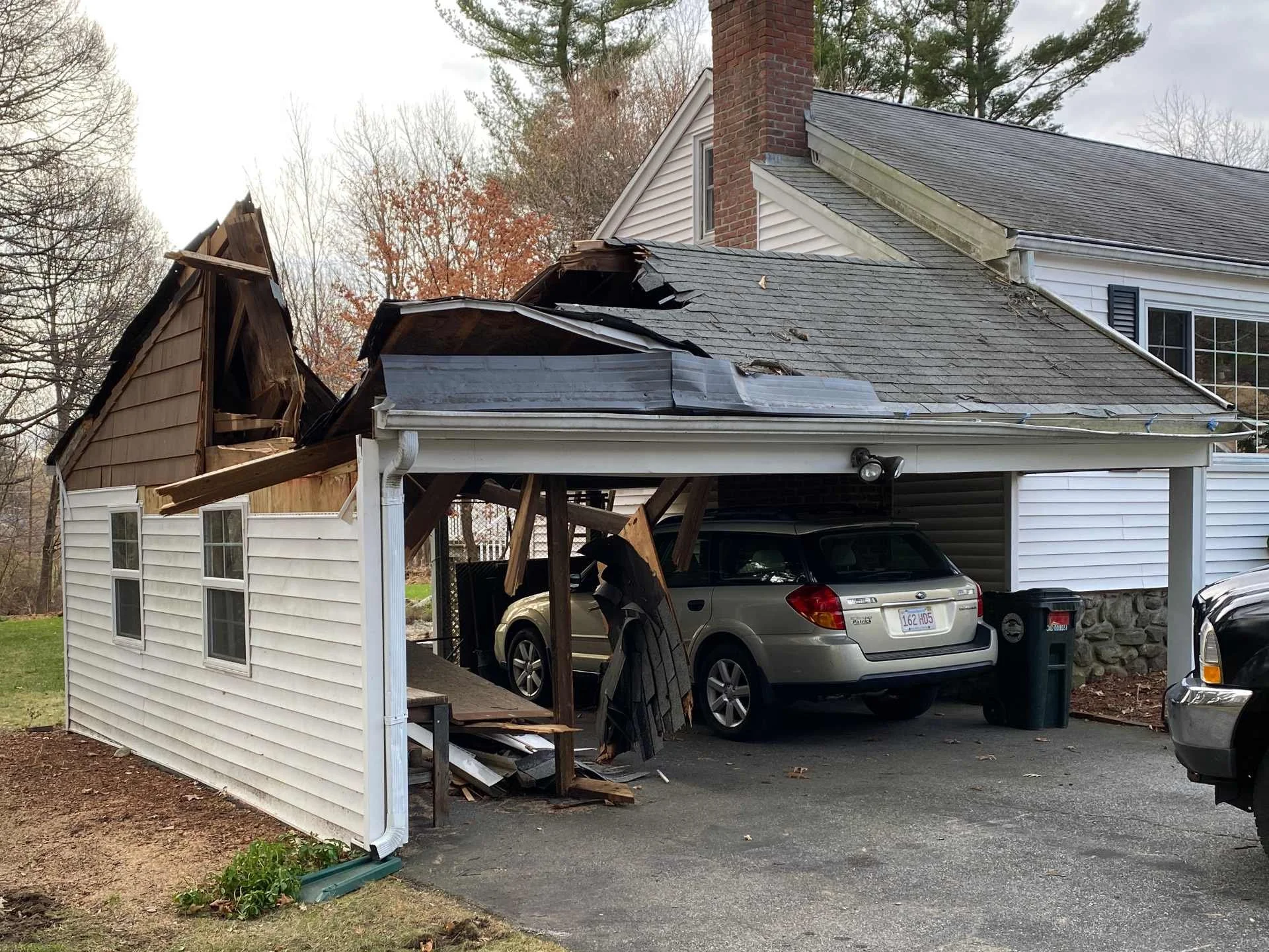 House with damaged roof and collapsed wall, debris on the driveway, cars parked under the carport, and trees in the background in Holden, MA.