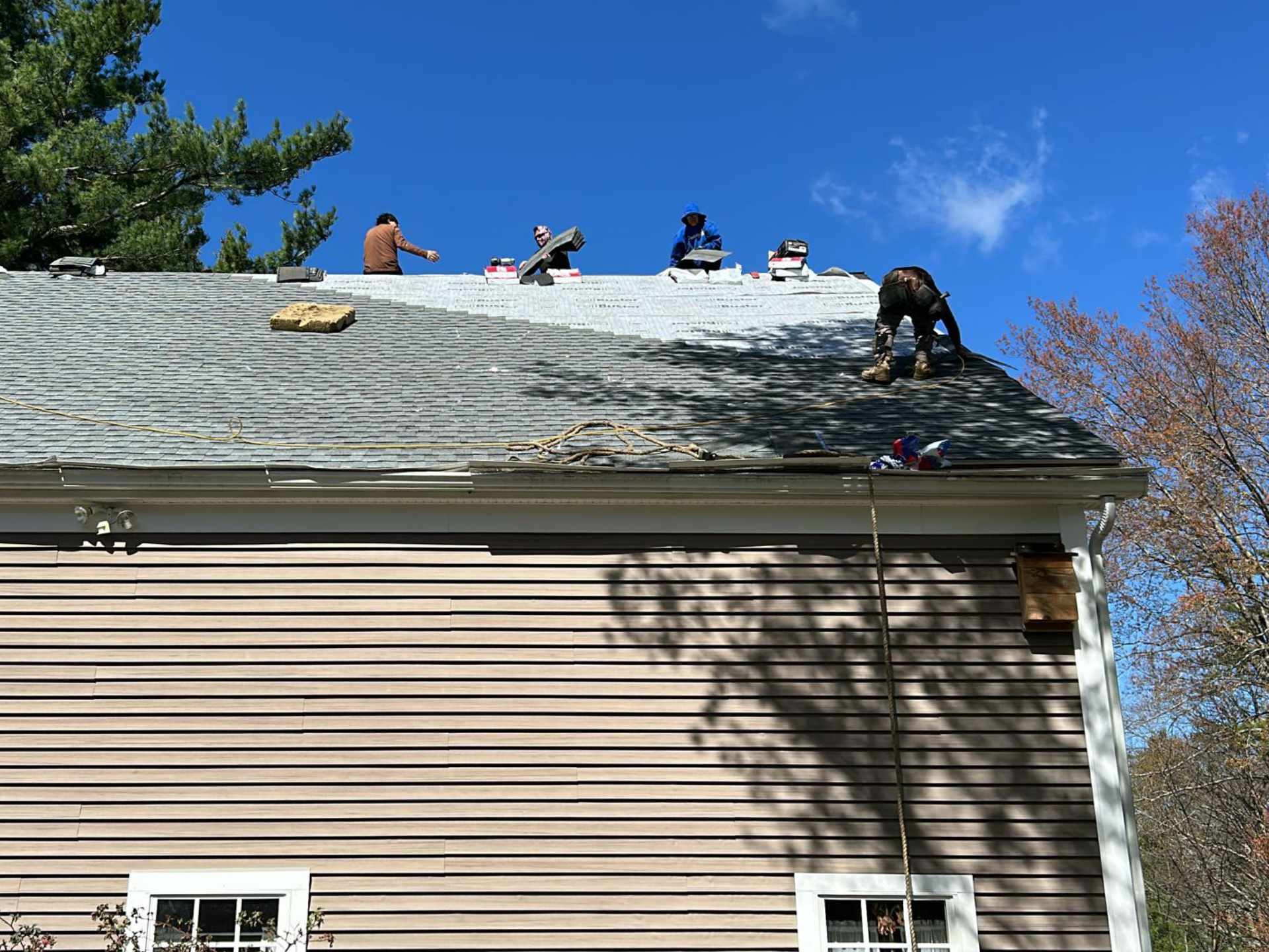 Workers on the roof installing new shingles in Winchendon, MA on a sunny day with a clear blue sky.