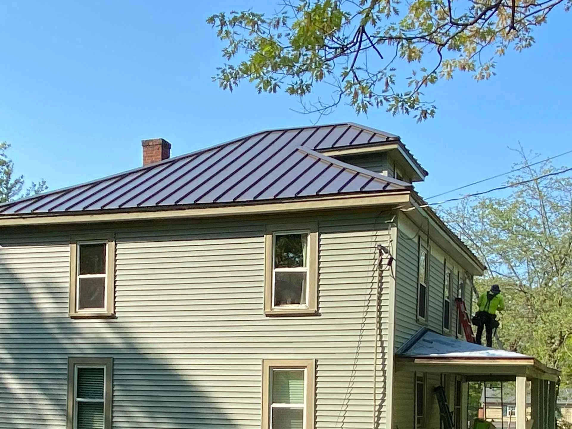 A two-story house with new seagrass green Certainteed Vinyl siding and a new Antique Bronze Standing-seam metal roof in Princeton, MA surrounded by green trees under a blue sky. Two workers are finishing the siding on the side of the house.