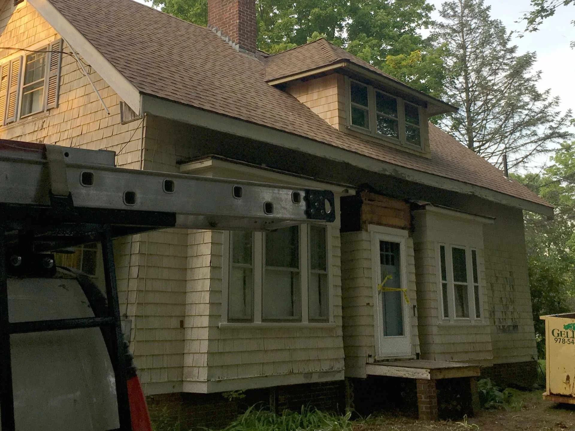 A house under renovation with a new front porch and windows being installed, construction equipment visible in the foreground, and trees in the background.